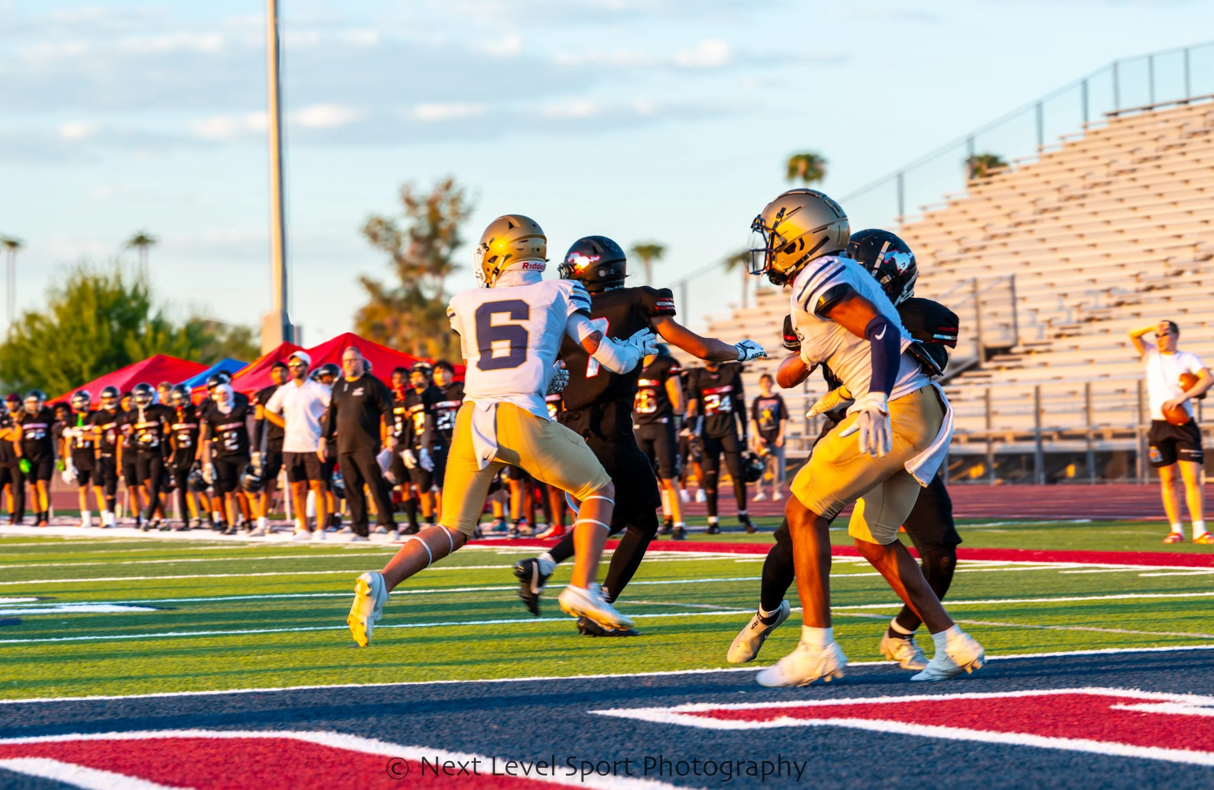 American football game with players on the field, one in black and three in white and yellow uniforms, during sunset, with coaches and other players in the background.