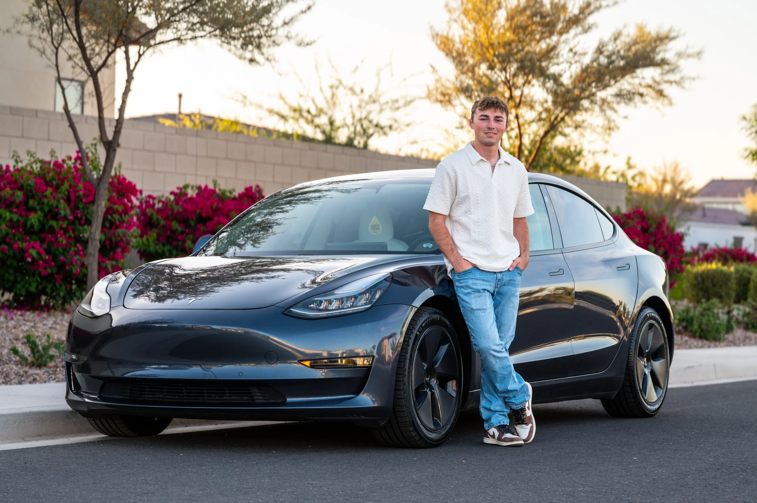 Young man leaning against a black Tesla car on a suburban street during sunset.
