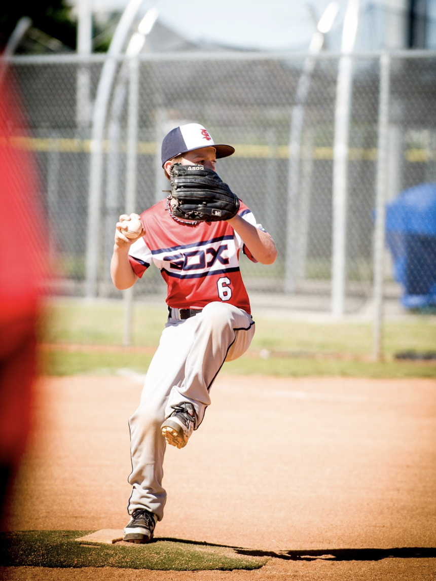 Youth baseball player in a red and white jersey with the number 6, wearing a cap and glove, pitching on a baseball field.