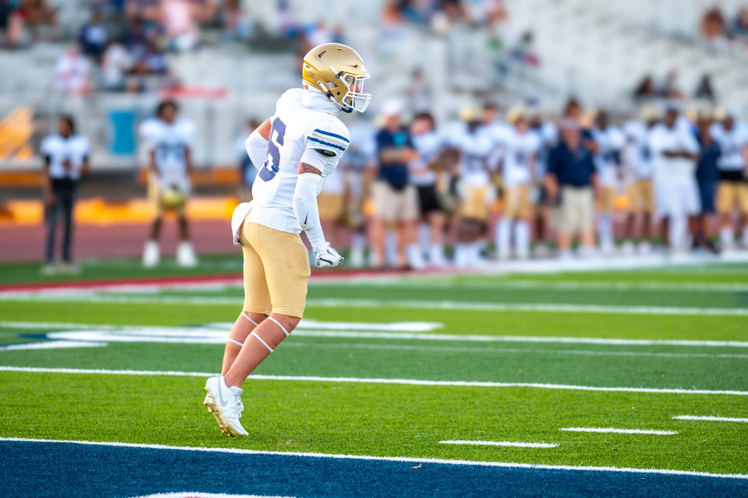 A football player in uniform and helmet standing on the field during a game, with teammates and spectators in the background.
