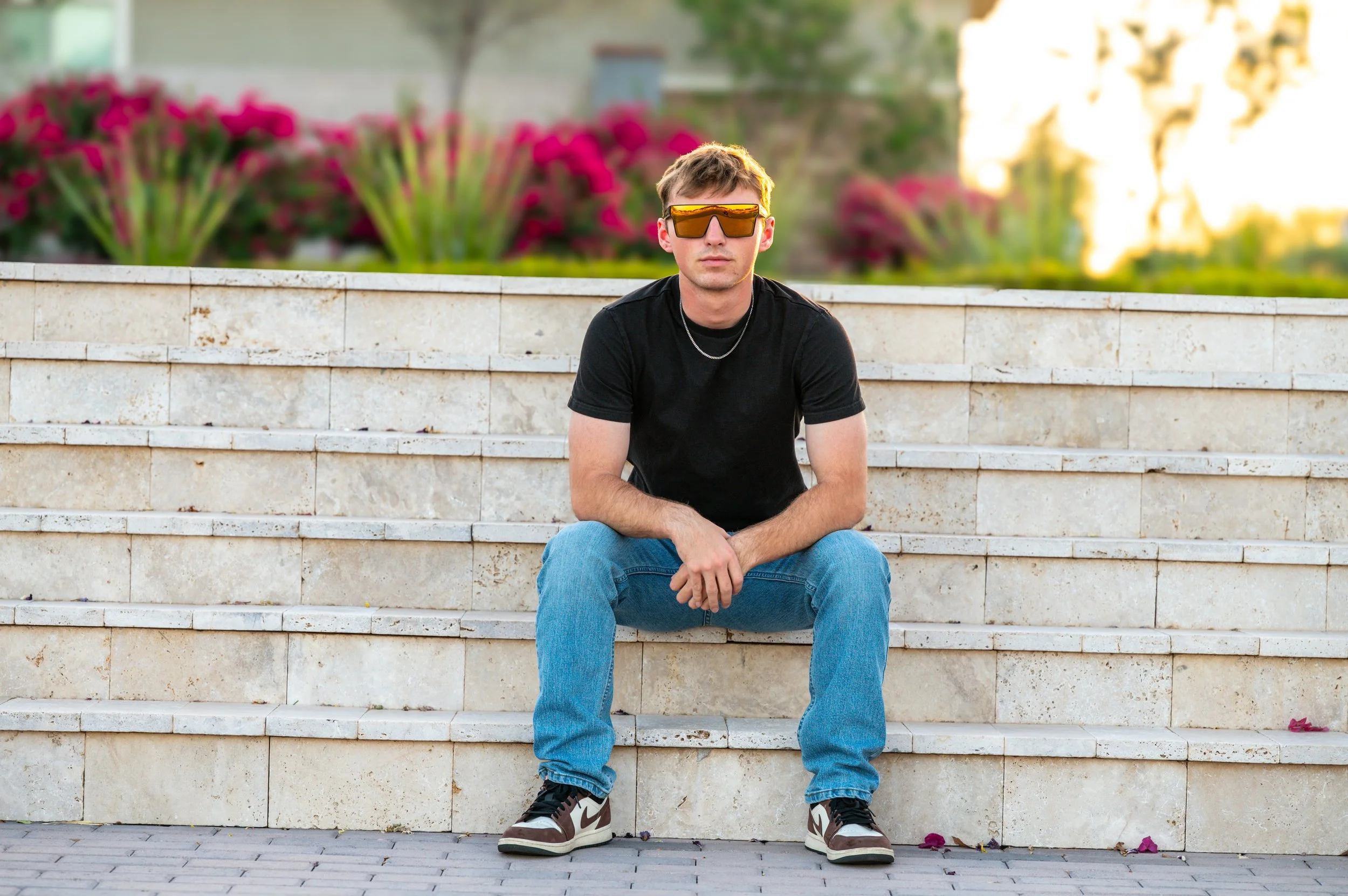 Young man sitting on stone steps outdoors, wearing a black t-shirt, blue jeans, sneakers, and orange sunglasses.
