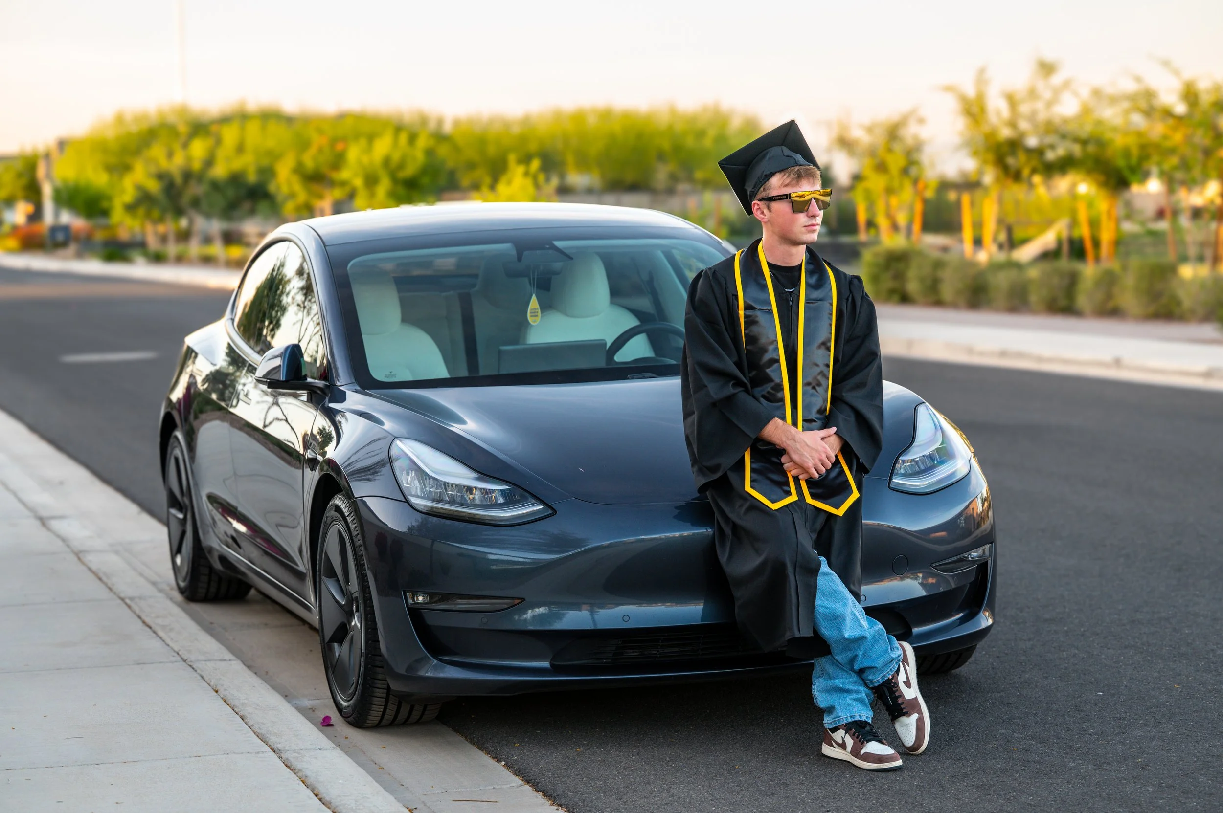 A young man in a black graduation gown and cap, wearing sunglasses, leaning against a black car on a street, with trees and a clear sky in the background.
