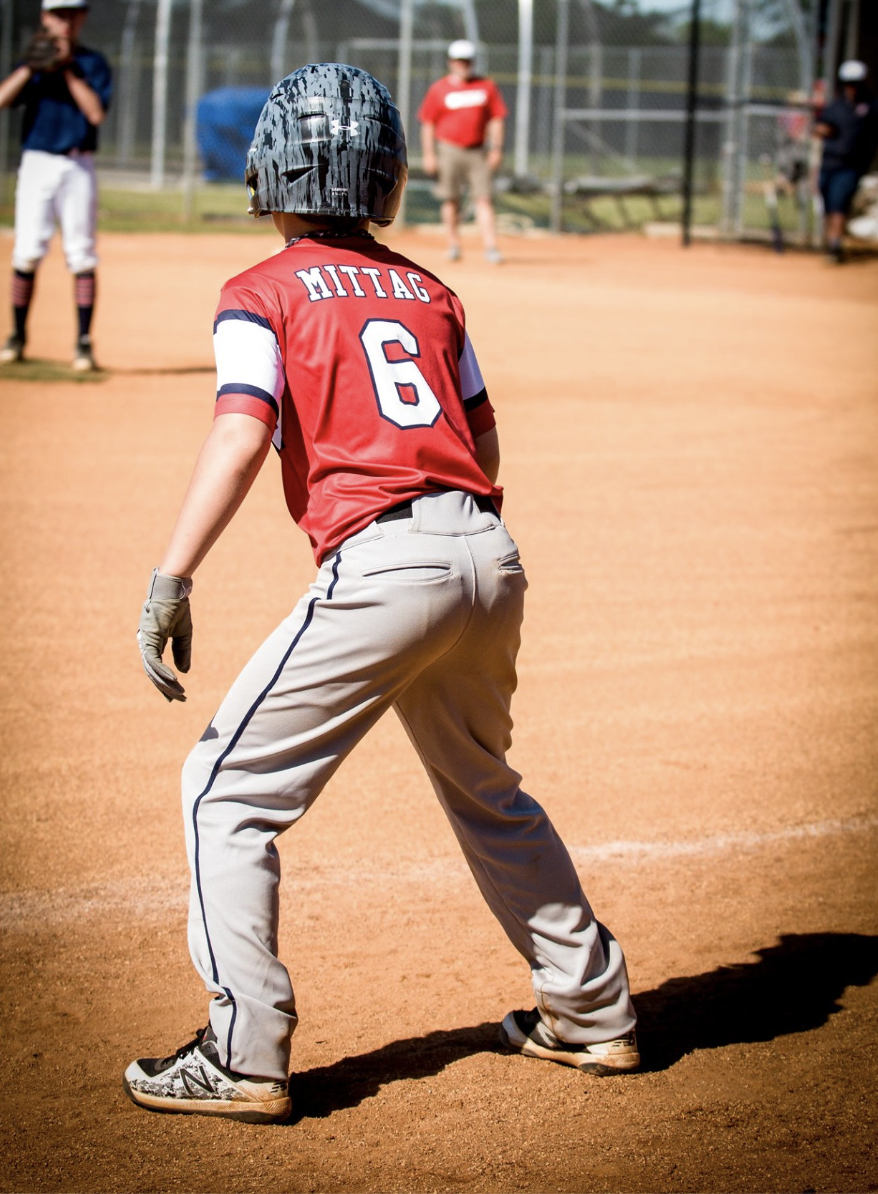 Young baseball player wearing a red jersey with the name Mittelg and number 6, standing on a baseball field in a ready stance, wearing a helmet and gloves, with other players and spectators in the background.