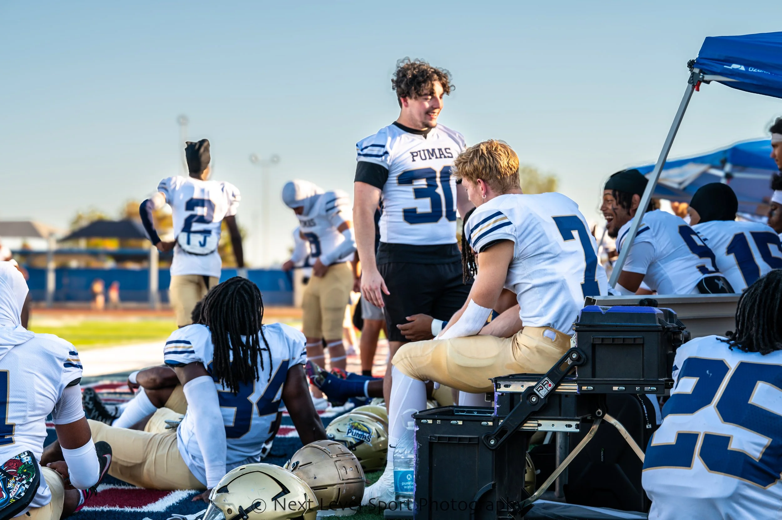 High school football team sitting on the sideline during a game, with players in white jerseys with blue and gold accents, some standing and some sitting on a bench, under a blue canopy, with helmets on the ground and a coach interacting with players