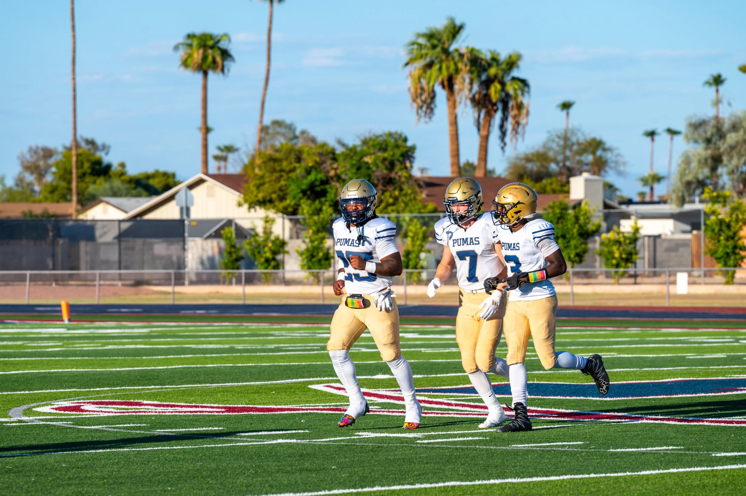 Three high school football players in uniform and helmets jogging on a football field during daytime with palm trees and houses in the background.