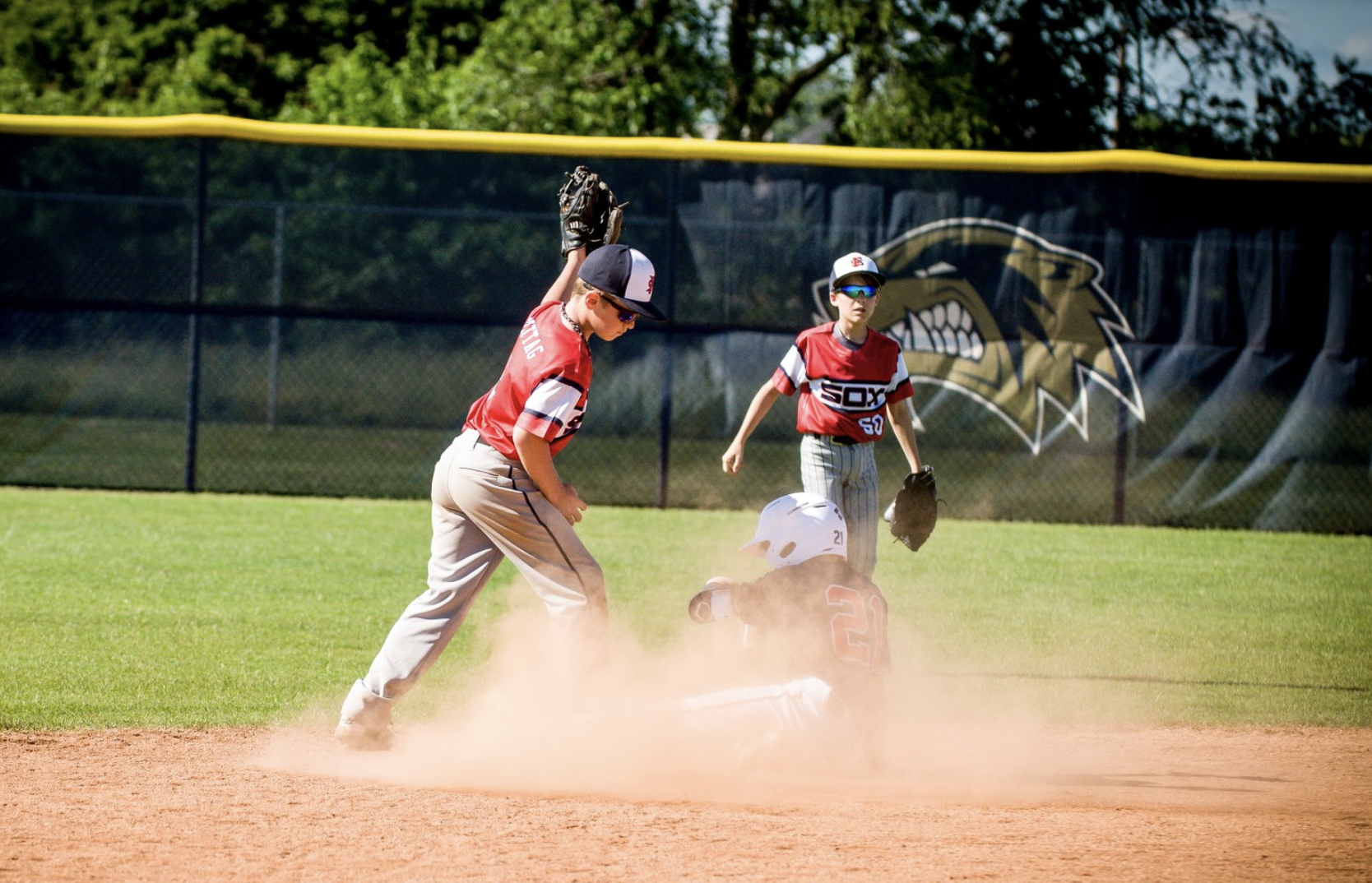 Young baseball players in red jerseys on the field, one sliding into second base, while a teammate and an opposing player watch nearby, with a fence and trees in the background.