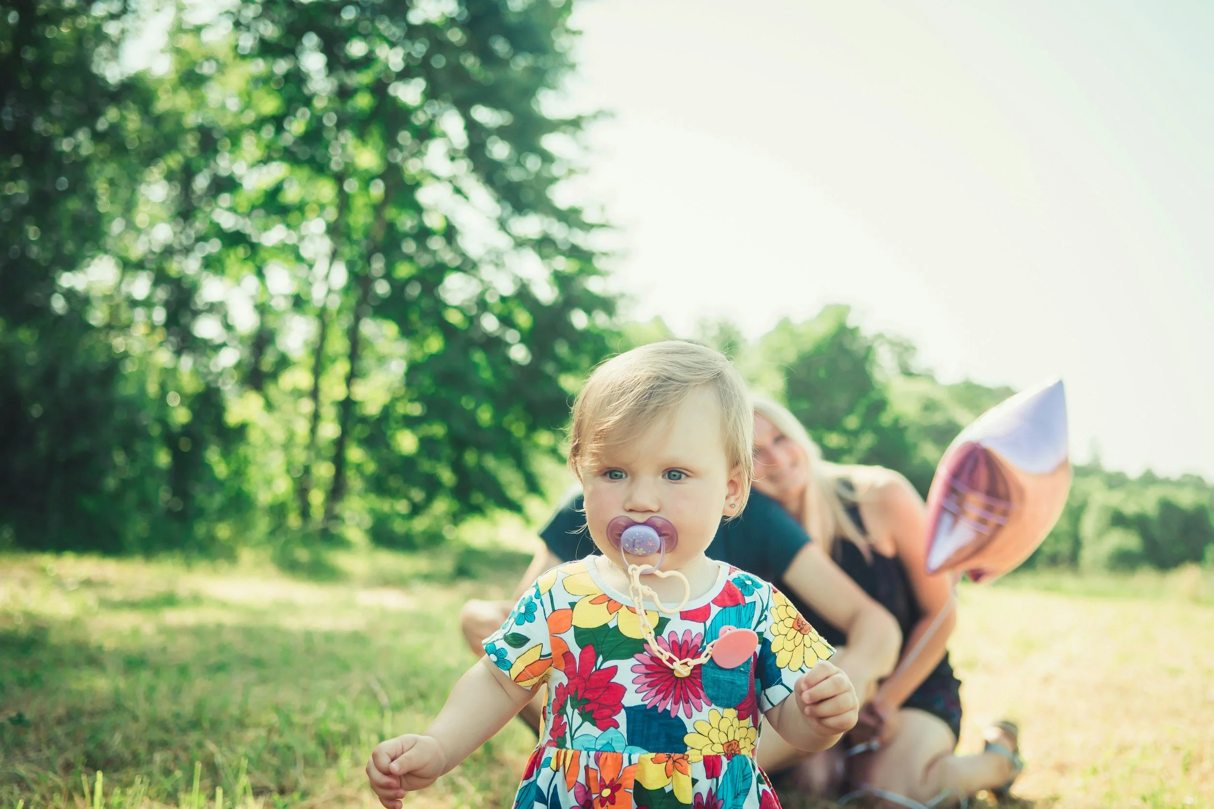 Young girl with blonde hair and a colorful floral dress walking with a pacifier in her mouth, a woman with blonde hair and a black outfit in the background outdoors on a sunny day, with trees and grass.