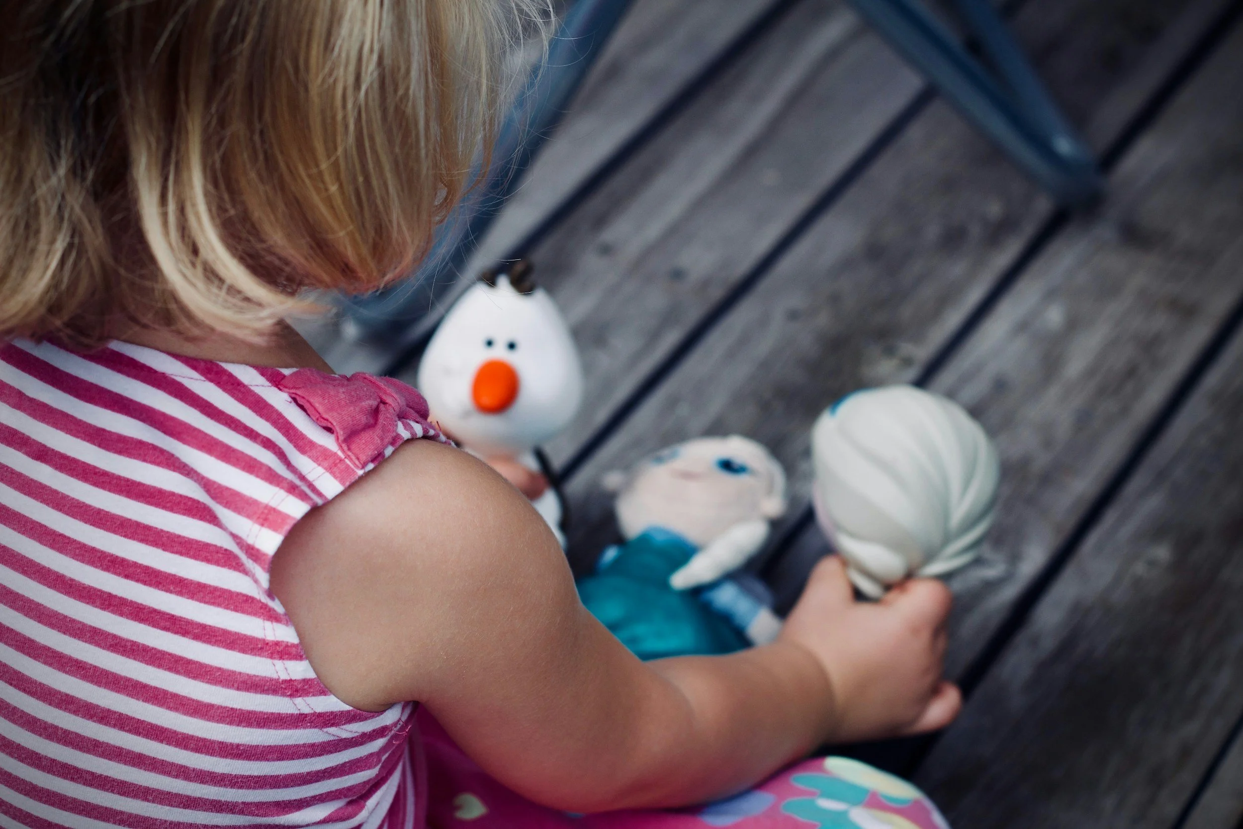 A young girl with blonde hair wearing a pink and white striped shirt kneeling on a wooden deck, holding a sphere-shaped toy with black and white stripes in her right hand, and with a stuffed doll and a white teapot with a snowman's face in front of her.
