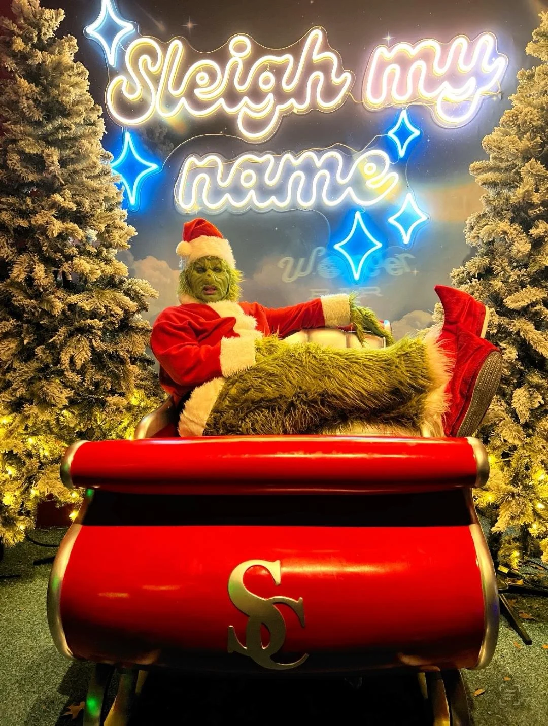 The Grinch dressed as Santa Claus sitting in a red sleigh with a neon sign behind him that says 'Sleigh my name'. There are two snow-covered Christmas trees on either side of the sleigh.