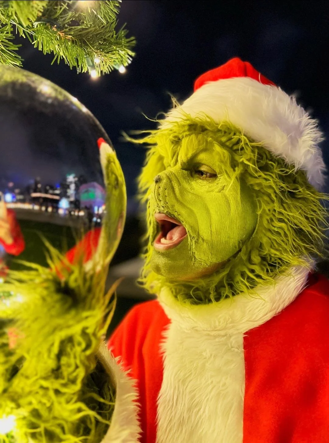 A person dressed as the Grinch with green face and furry gloves, wearing a red Santa outfit and Santa hat, holding a shiny Christmas ornament.