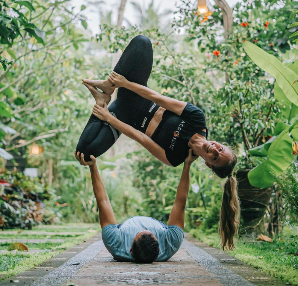 Two people practicing acro yoga outdoors on a garden path, with the person on the ground supporting the person lifted above them. The person on the ground is lying on their back with their legs bent and arms raised to hold the other's arms. The person lifted is in a balancing pose, smiling, with their body angled sideways and one leg bent, surrounded by lush green foliage and plants.