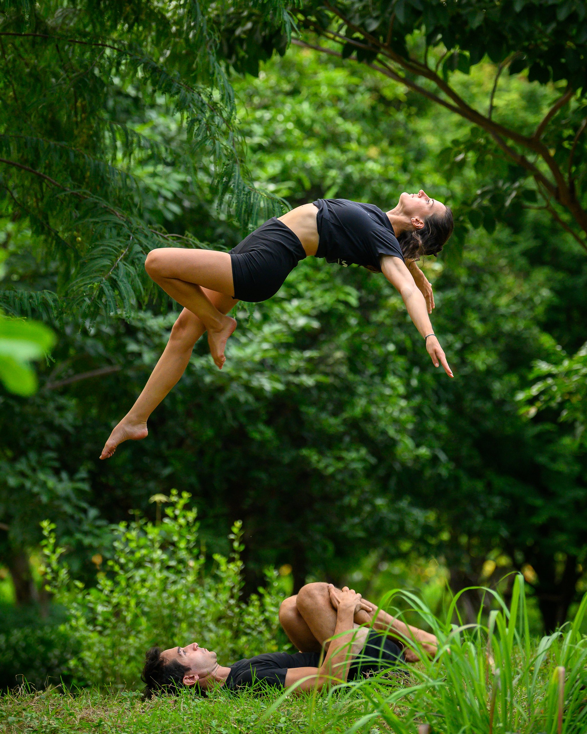 Two people practicing acro yoga outdoors in a lush green forest. One person is lying on their back on the grass, holding their knees close to their chest, while the other is balanced on top, extended with arms outstretched.