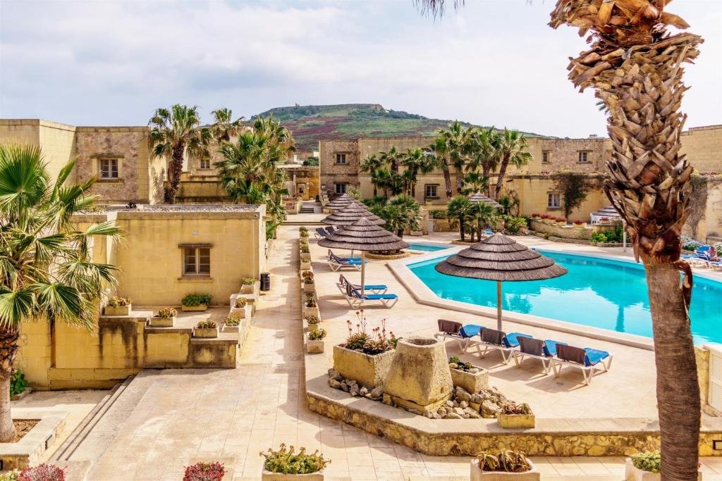 Luxury resort pool area with palm trees, thatched umbrellas, lounge chairs, and historic stone buildings, with a hill in the background.