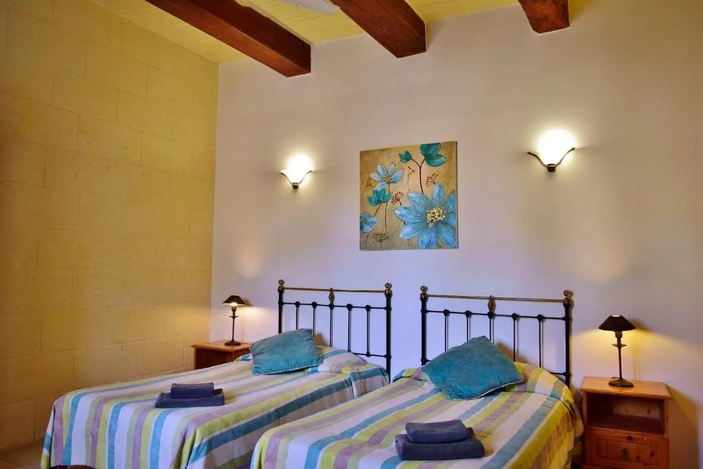 A bedroom with two twin beds, each with colorful striped bedding and blue pillows, wooden nightstands with small lamps, a floral painting on the white wall, and wooden ceiling beams.