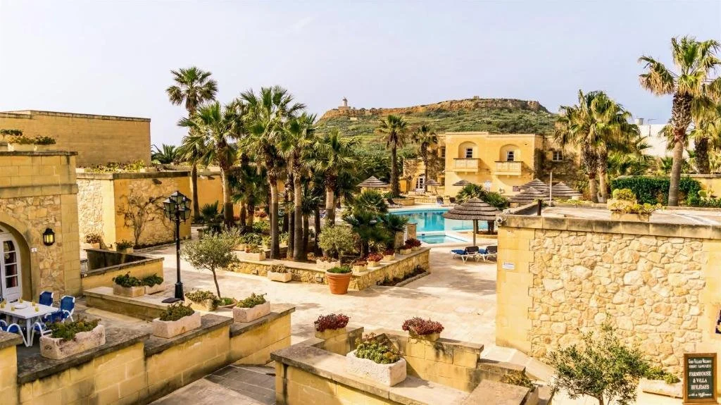 Resort with palm trees, stone buildings, a swimming pool, lounge chairs, and umbrellas in a sunny location with a hill in the background.