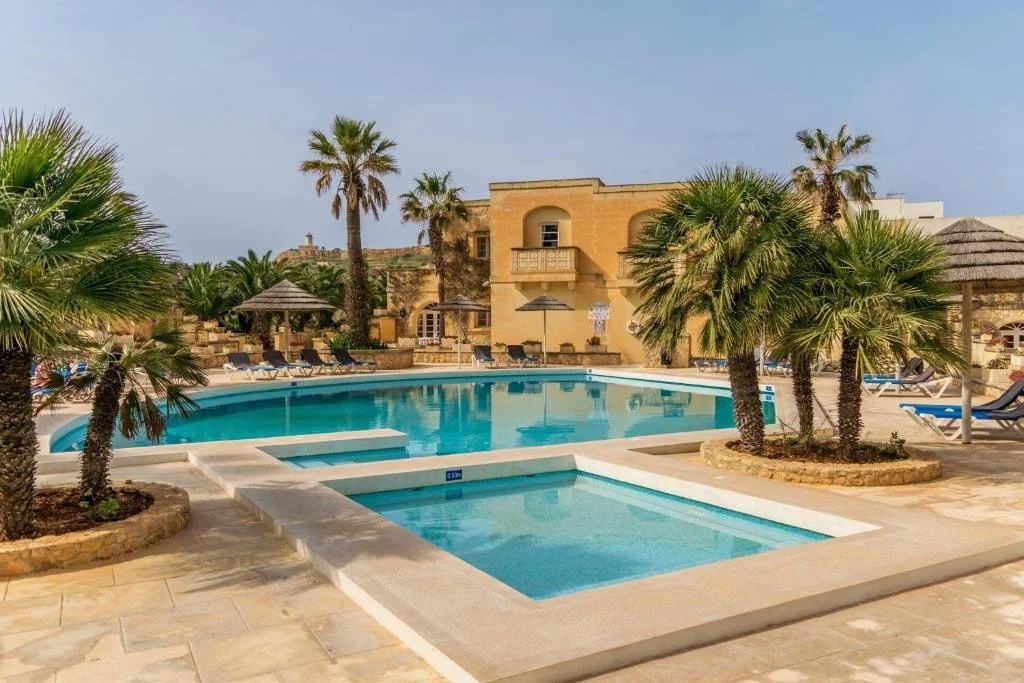 A resort pool area with a large swimming pool, surrounding palm trees, lounge chairs, and a building in the background, under a partly cloudy sky.