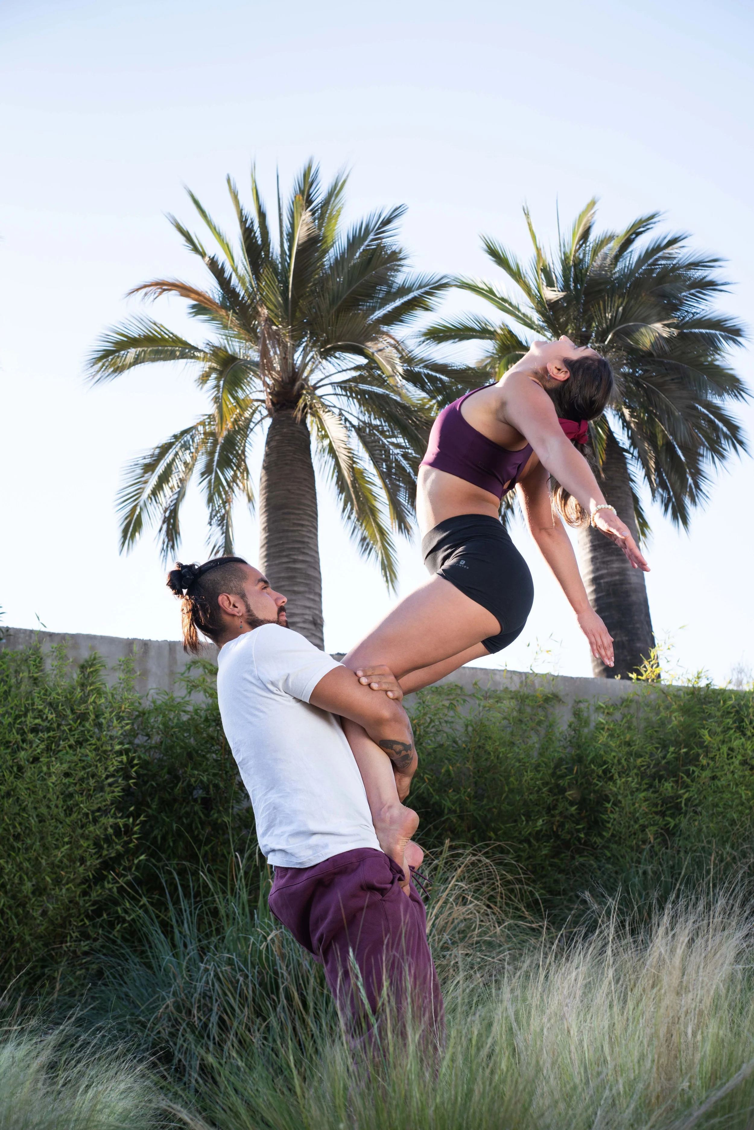 A man is balancing a woman on his shoulders while she stretches, outdoors with palm trees and greenery in the background.