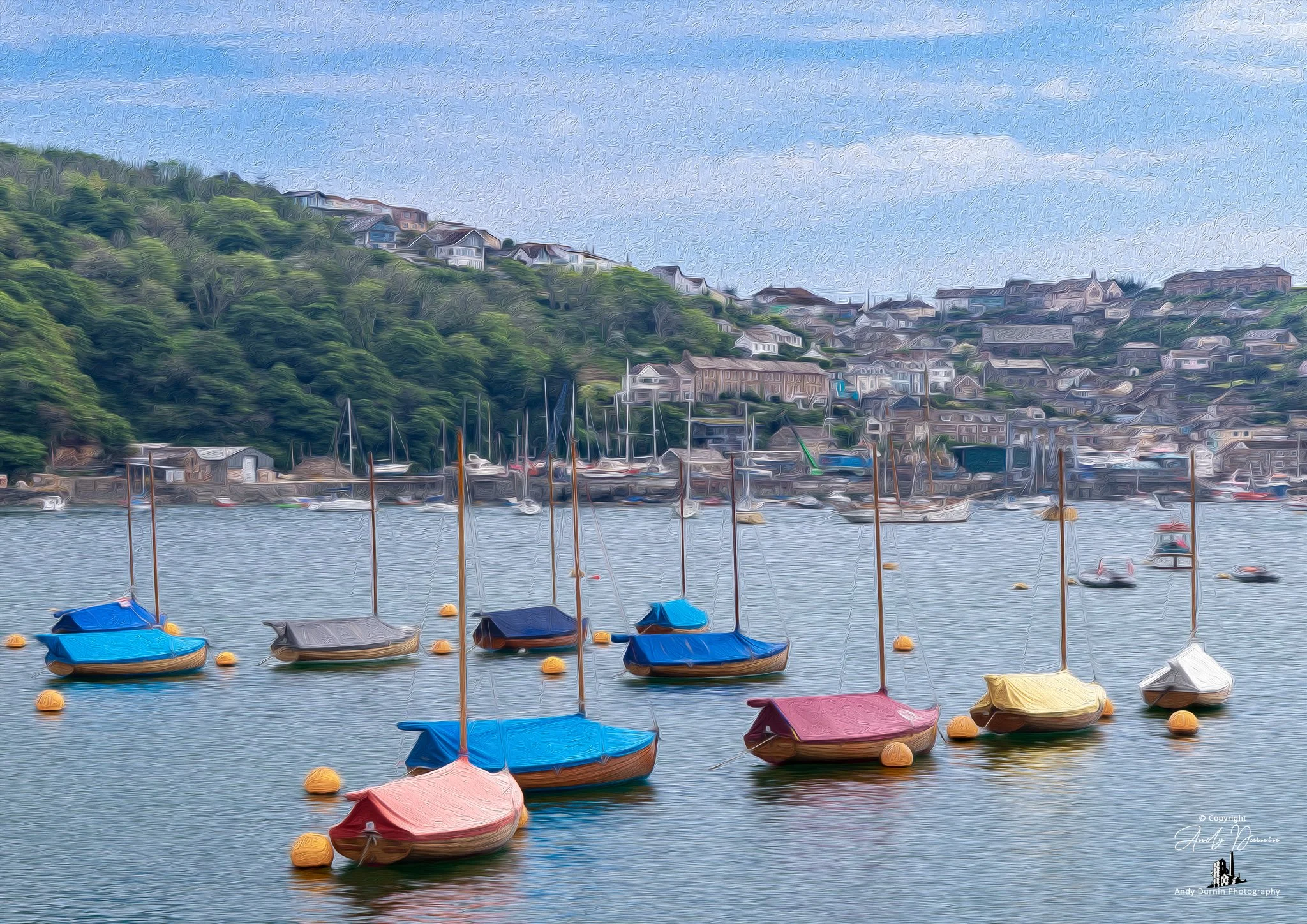The River Fowey and sailing dinghies  anchored in a harbour with colourful covers, with a hilly coastal town and houses in the background and a cloudy sky.