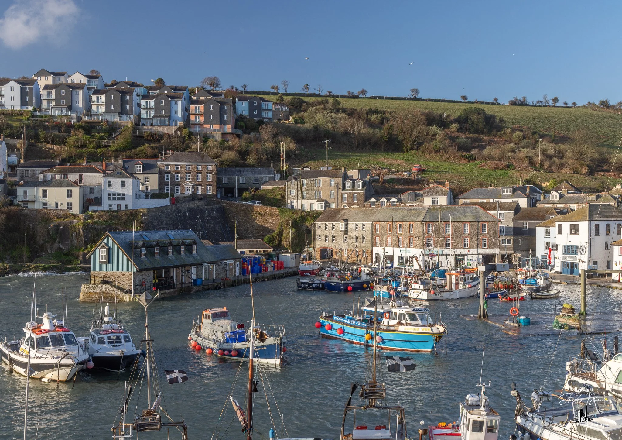 Fishing boats navigating Mevagissey Harbour on a bright, breezy day, with traditional stone buildings and hillside cottages rising above the busy waterfront.