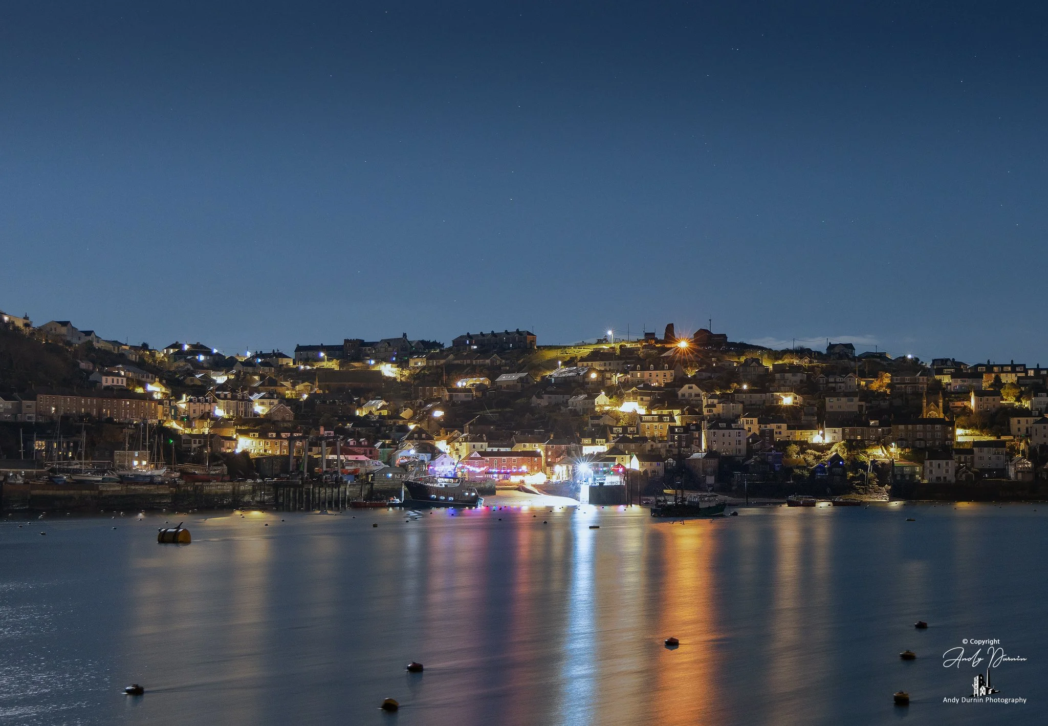Night-time long exposure of Polruan, with warm harbour lights reflecting across calm water under a deep, star-speckled sky. A moody Cornwall night print that captures quiet coastal atmosphere, glowing colour, and the serenity of the estuary after dar