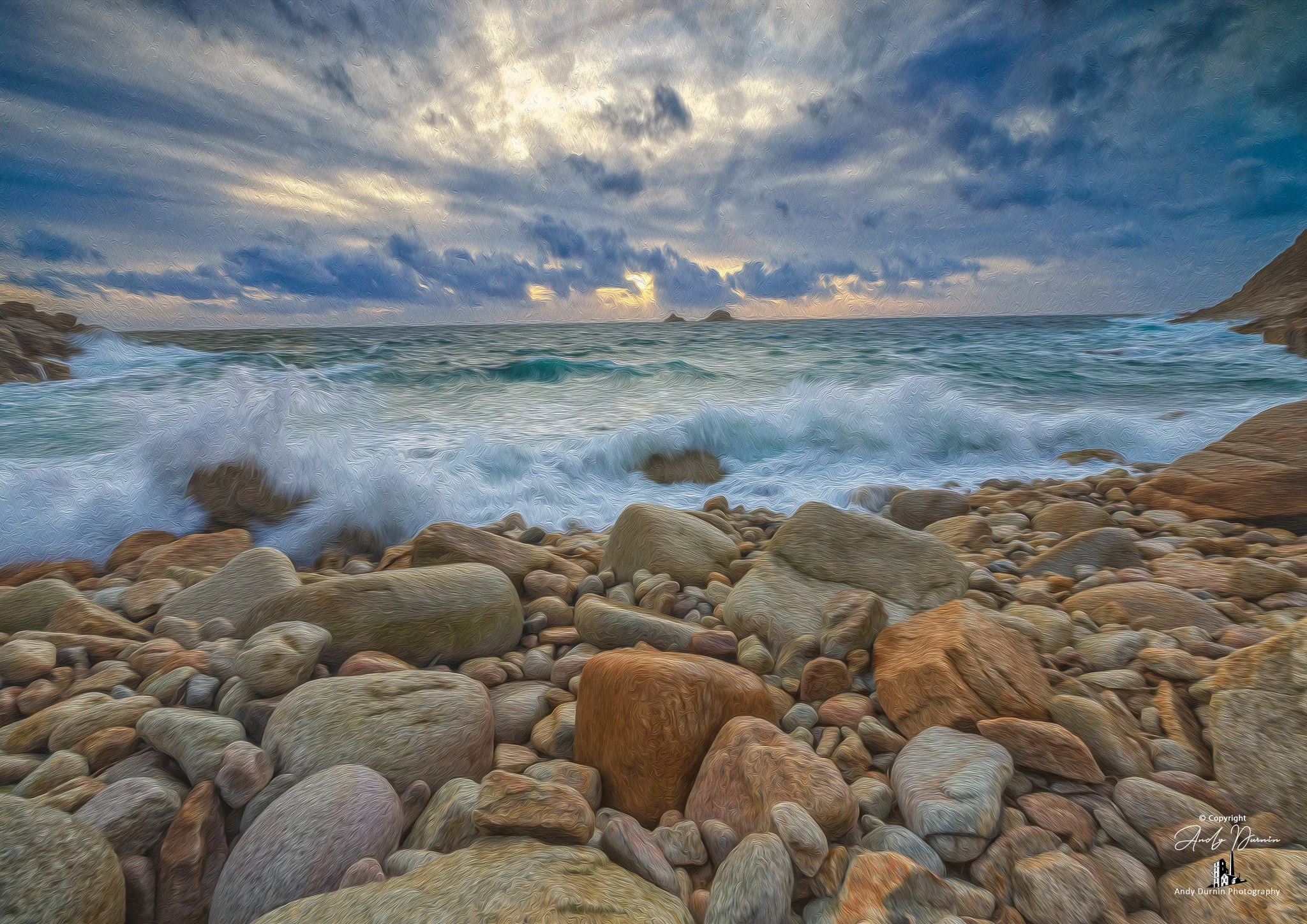 Porth Nanven (the Cot Valley) A rocky beach with large stones in the foreground and waves crashing against the rocks. The ocean extends to the horizon with a cloudy sky overhead, showing some sunlight breaking through.