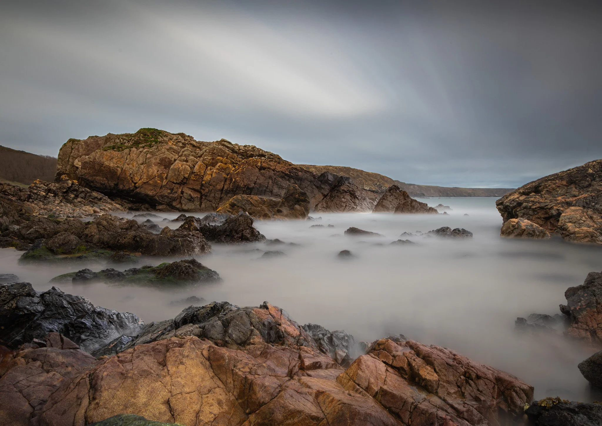 A rocky coastline with large boulders and rocks in the foreground, a large rock formation on the left, and a calm ocean with misty water effects. Overcast sky with moving clouds.