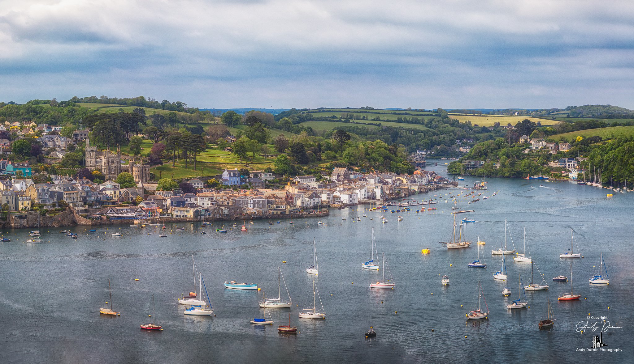 A wide panoramic view of Fowey on the River Fowey, Cornwall, with colourful waterfront buildings, moored boats and rolling green hills under dramatic skies—ideal coastal wall art.