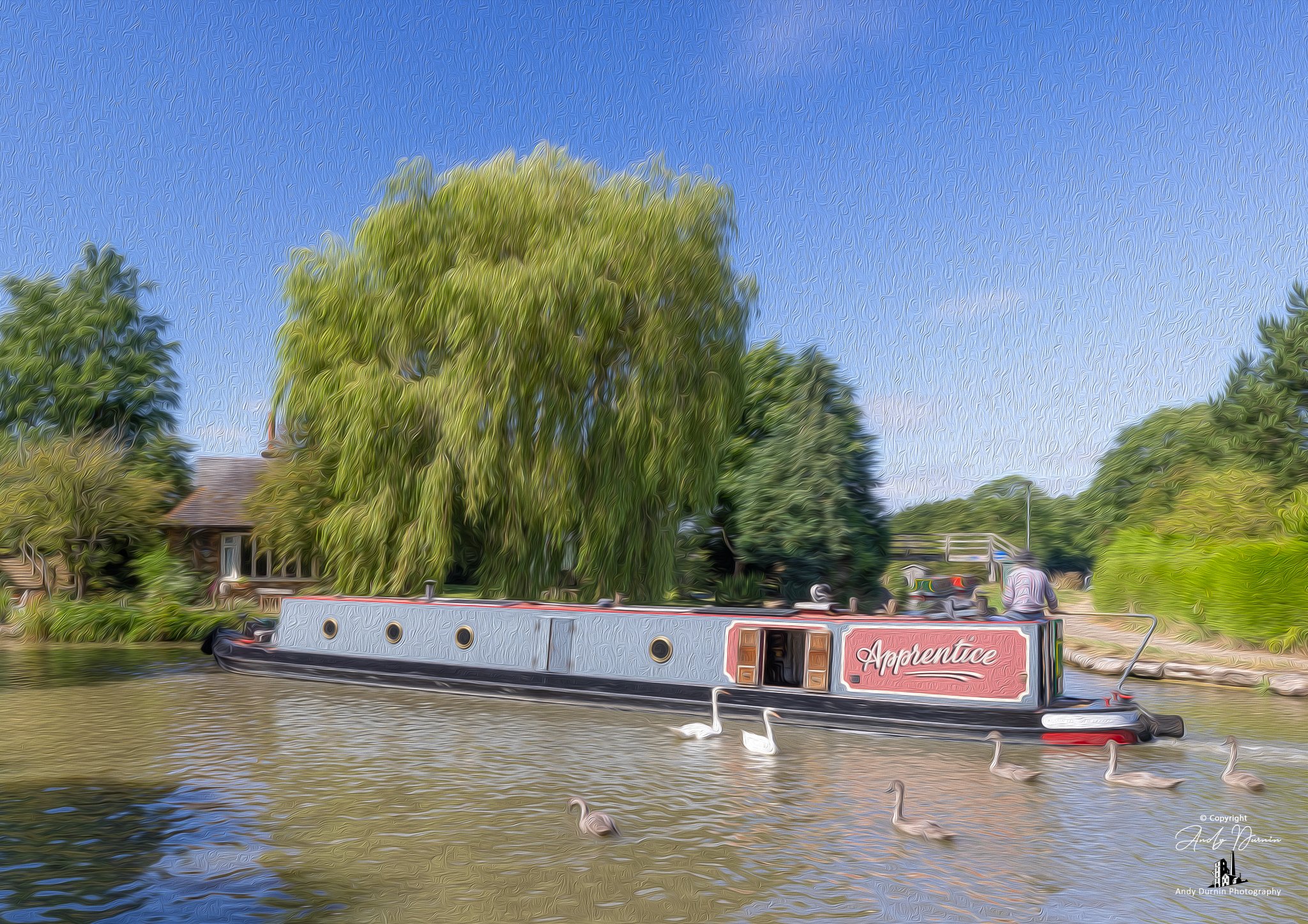 Norton Junction on the Grand Union Canal.  A canal scene featuring a narrowboat called the  'Apprentice' , surrounded by trees, ducks swimming in the water, and a person sitting on the boat's roof under a sunny, blue sky.