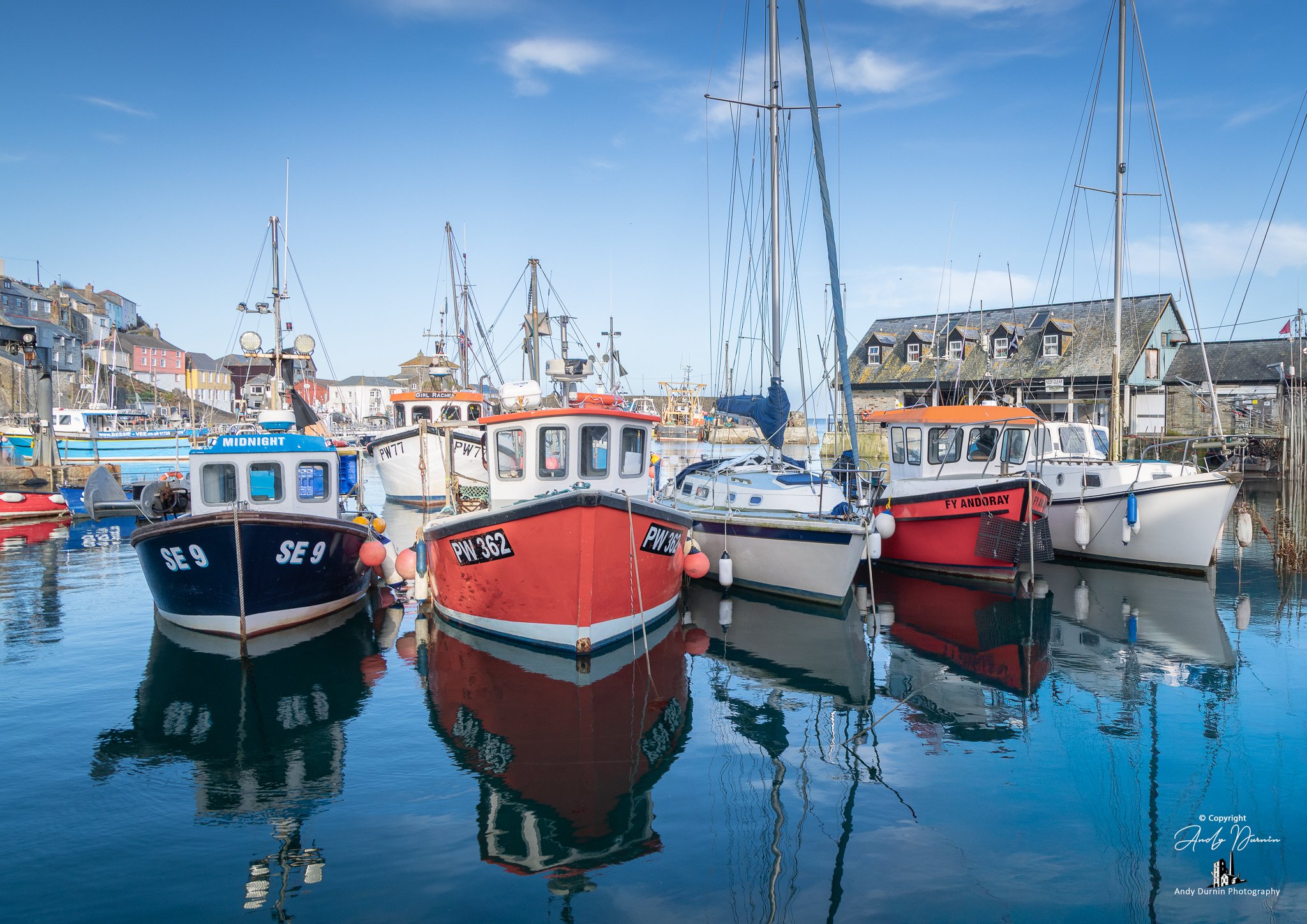 Fishing boats moored side by side in Mevagissey Harbour on a calm, clear day, with crisp reflections in the water and bright blue skies above.