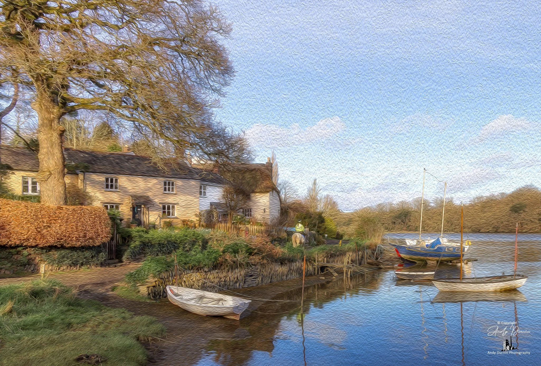 St Clement on the River Tresillian a peaceful riverside with houses, trees, and boats docked along the water's edge under a blue sky with a few clouds.