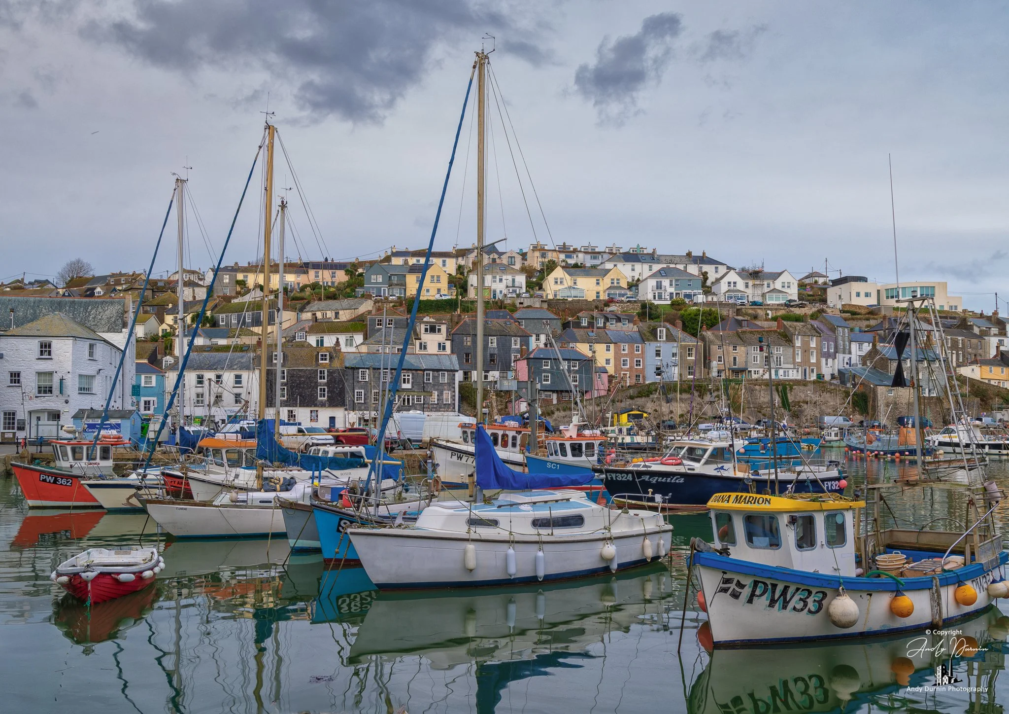 Colourful boats moored in Mevagissey Harbour with rows of pastel cottages and traditional houses rising up the hillside under a cloudy coastal sky.