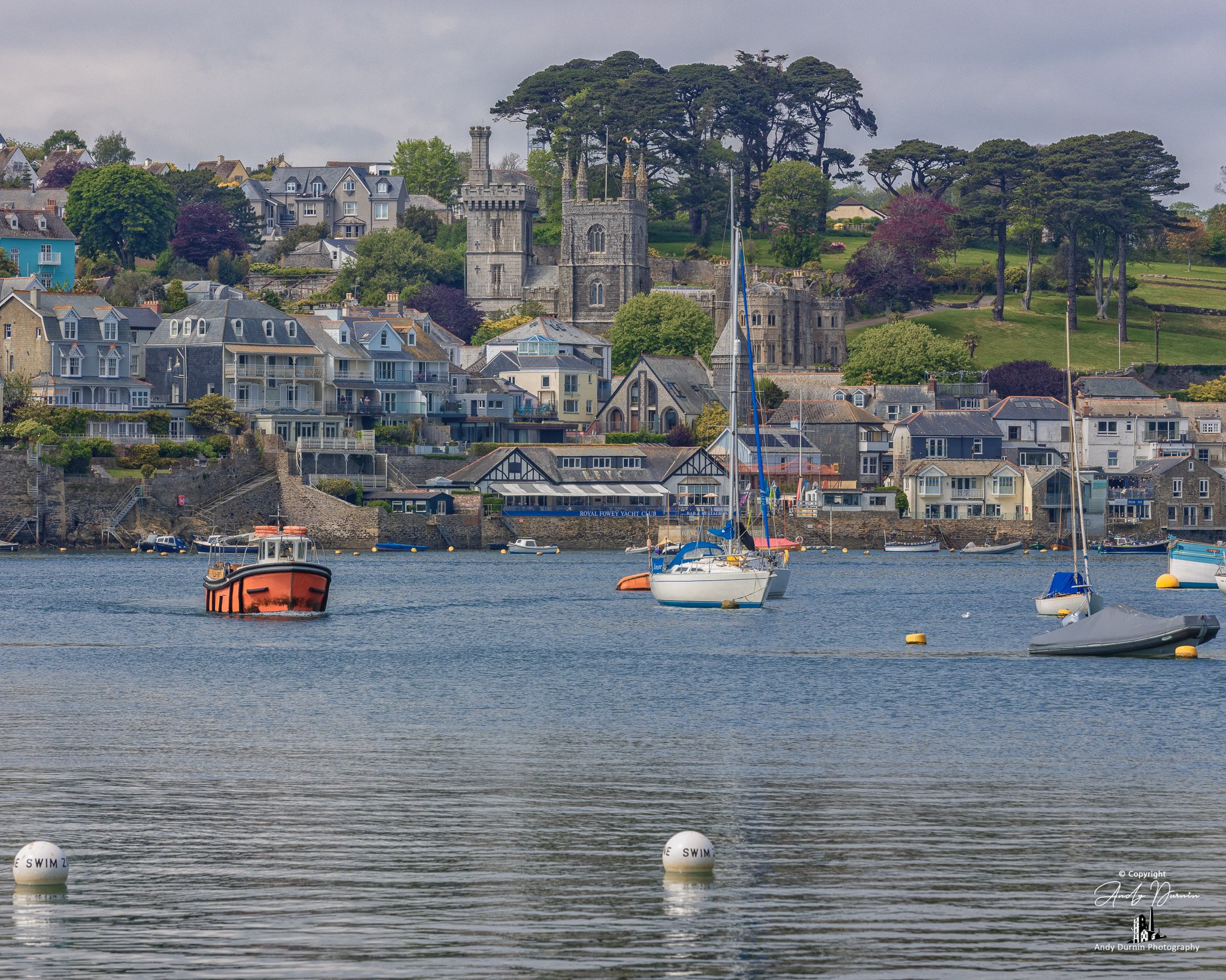 A detailed riverside scene of Fowey, Cornwall featuring the Church of St Fimbarrus with Place House behind, framed by boats on the River Fowey—classic Cornish harbour-town photography.