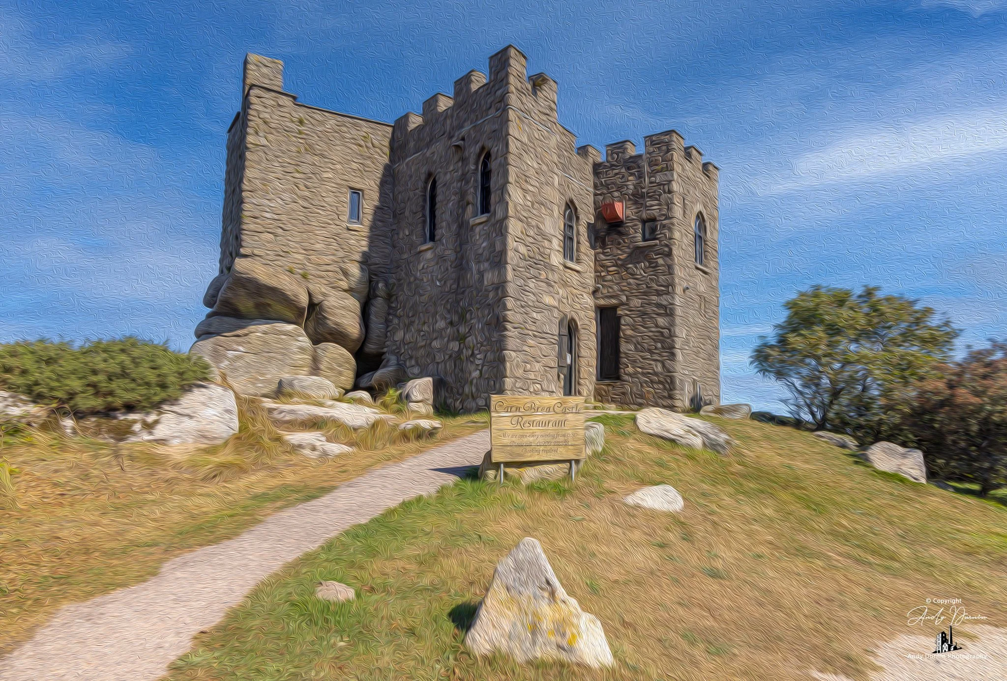 Carn Brea Castle.  A stone castle on a grassy hill with a dirt path leading up to it under a blue sky with scattered clouds.