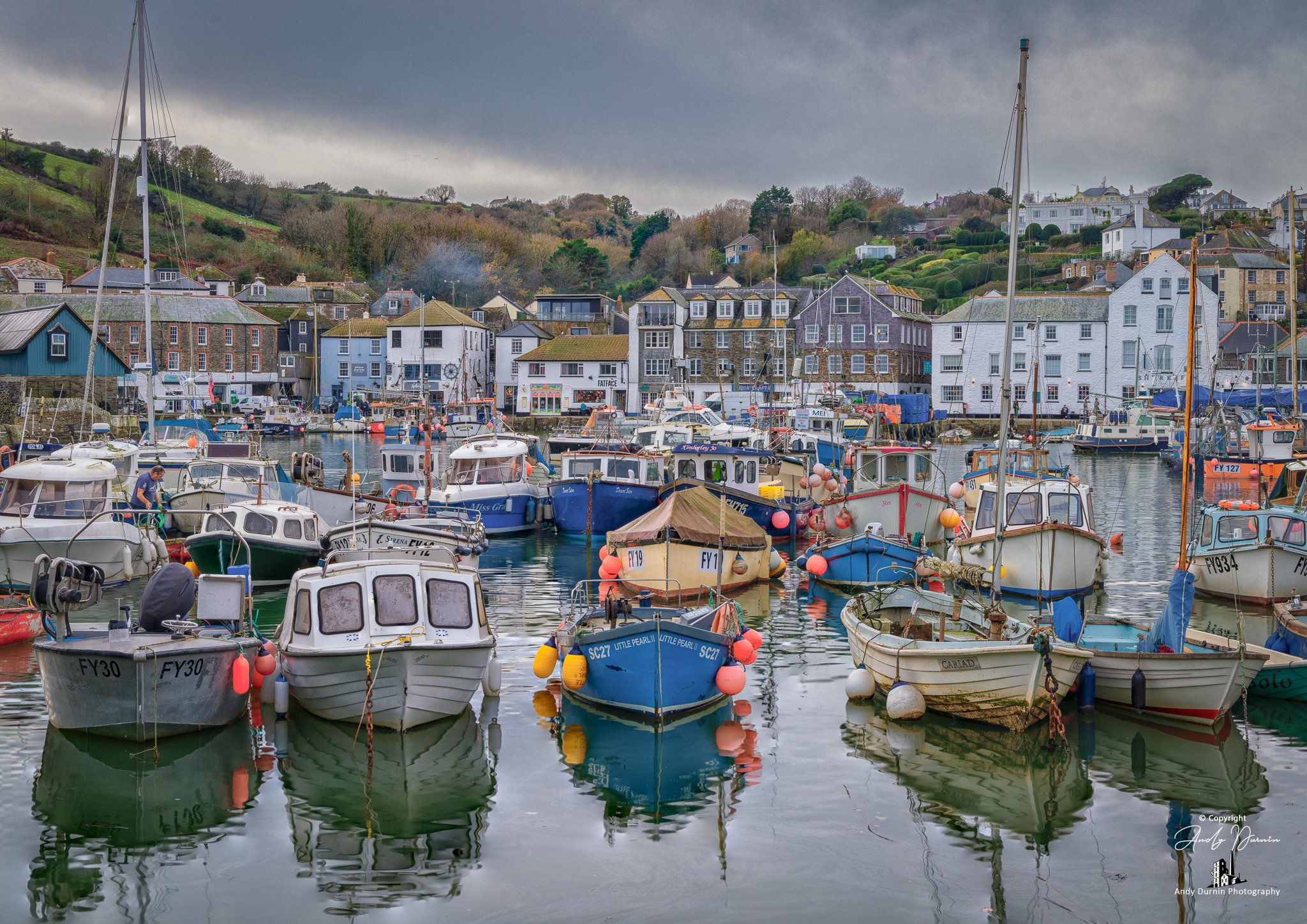 A busy view of Mevagissey Inner Harbour filled with colourful fishing boats, calm water reflections and traditional Cornish cottages rising up the hillside behind.