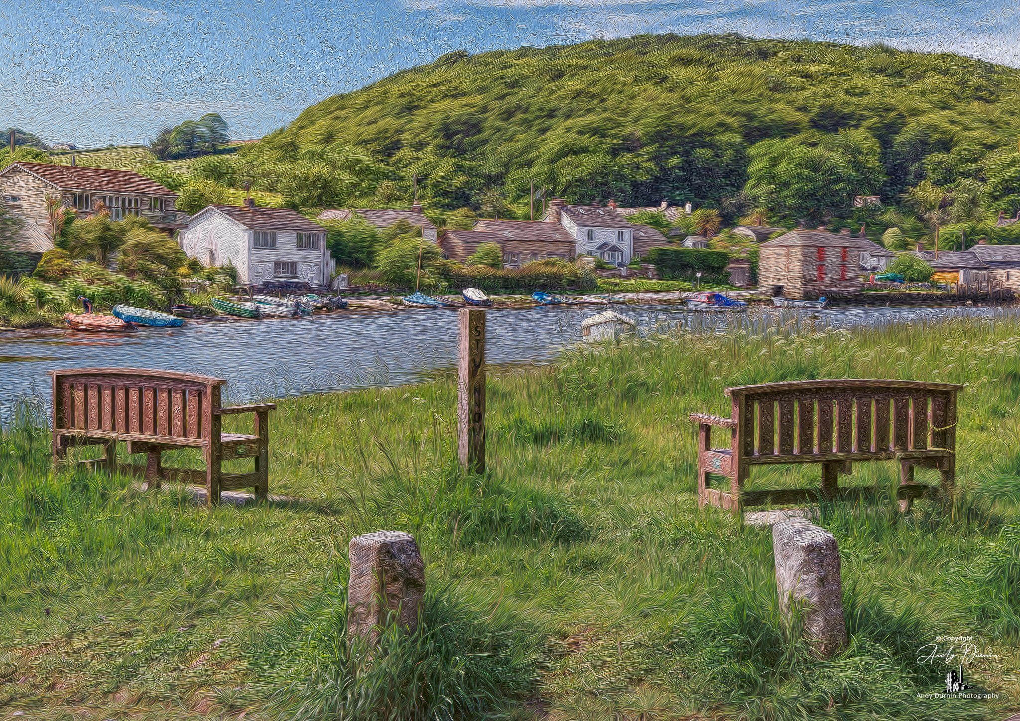The River Lerryn Cornwall Scenic view of a small waterfront village with houses and boats, two wooden benches on a grassy area in the foreground.