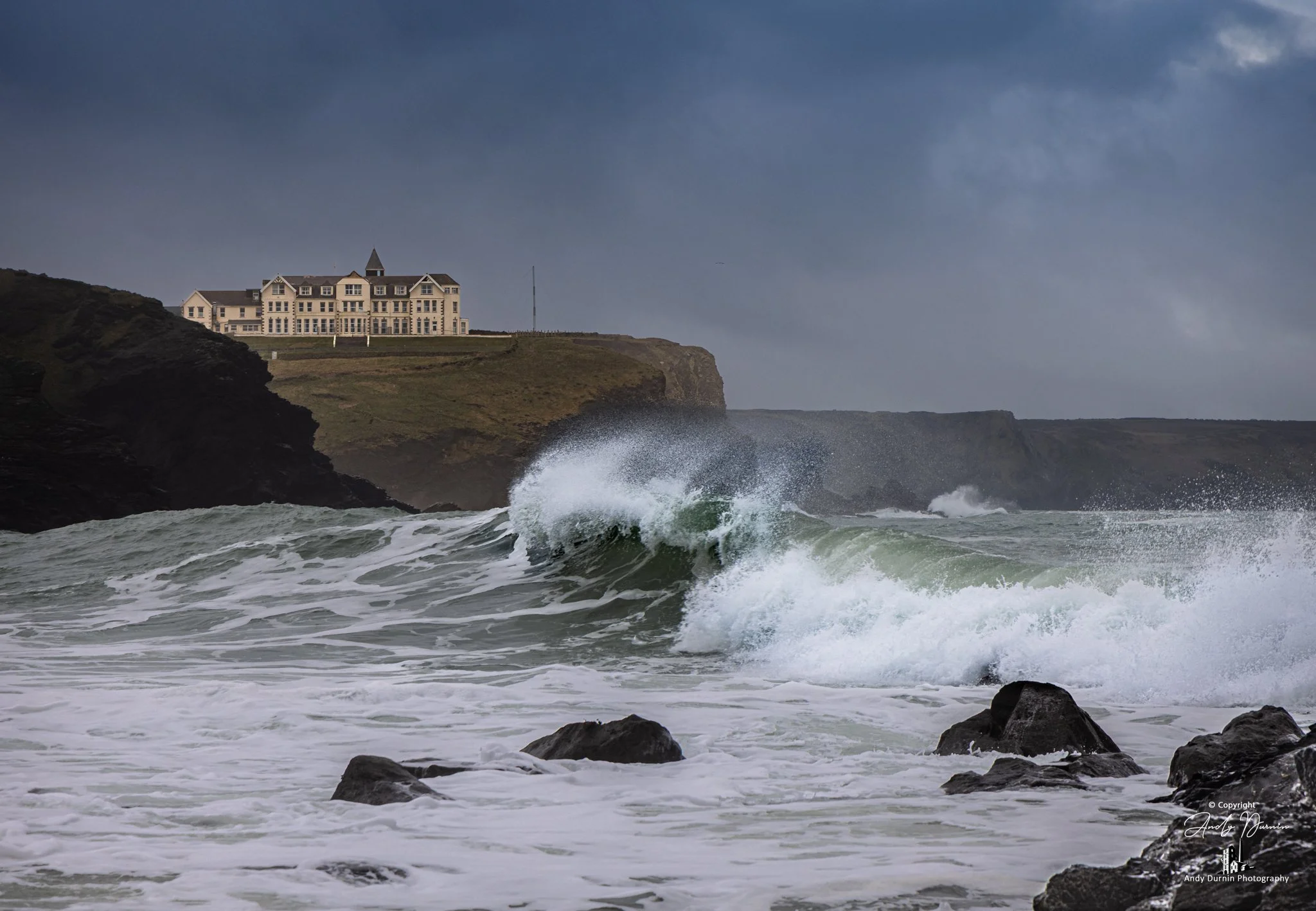 Moody storm waves crash into Church Cove, Gunwalloe, with Poldhu Nursing Home on the headland. Dramatic Cornwall seascape print by Andy Durnin.