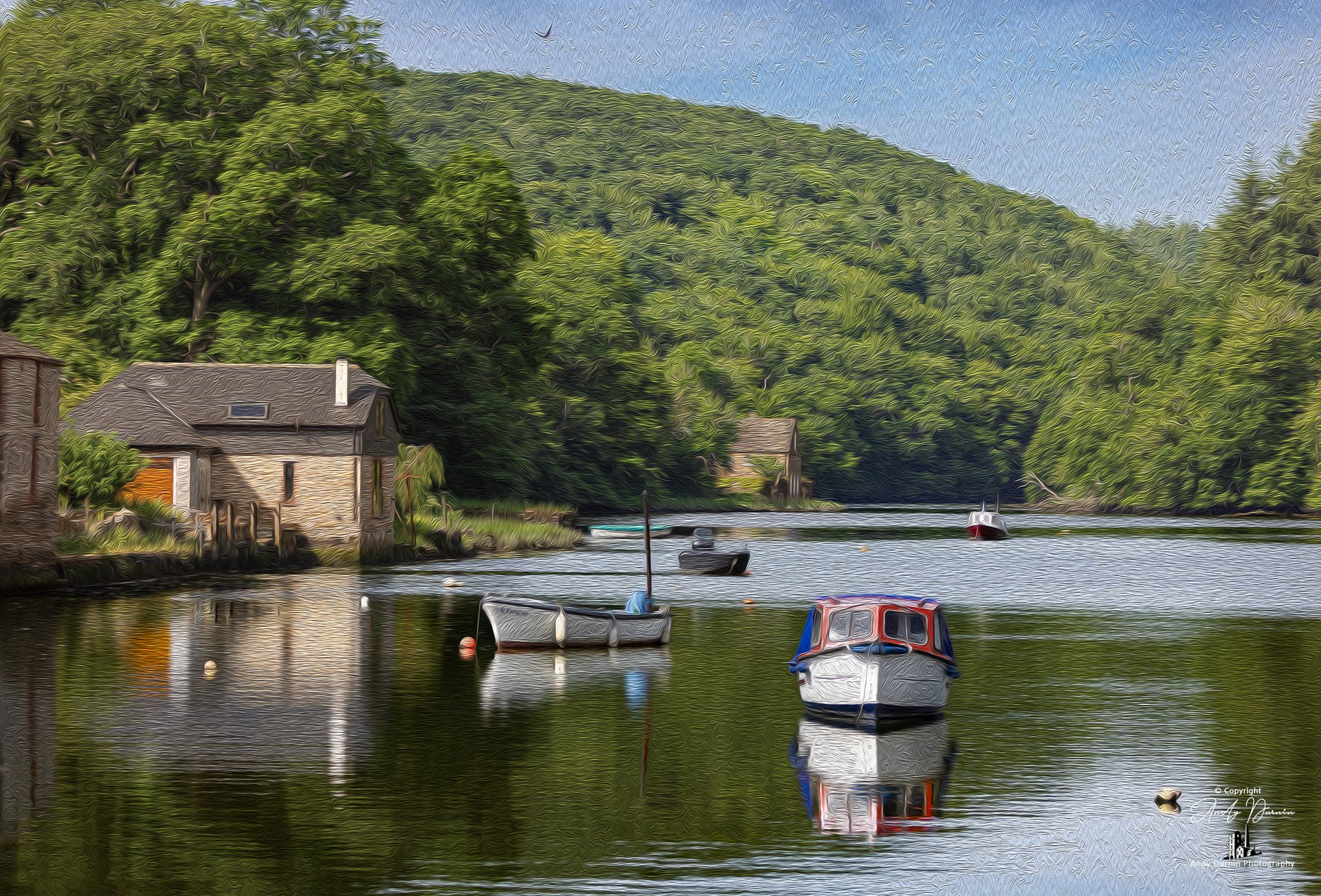 The River Lerryn Cornwall A scenic view of a river with boats docked and small houses along the shore, surrounded by lush green hills under a clear sky.