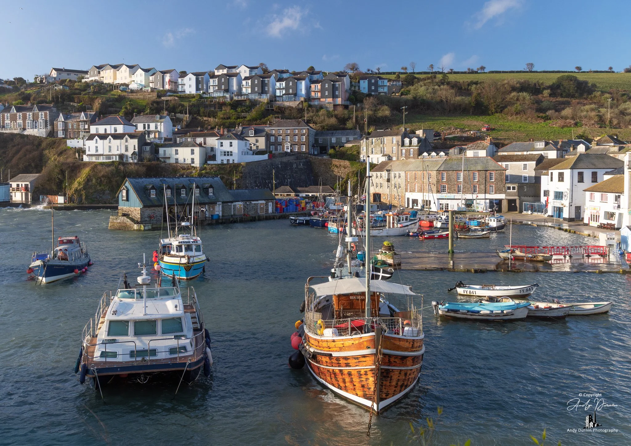 Fishing boats entering Mevagissey Harbour on a bright coastal day, including  the boat Seascan on the right, with hillside cottages and historic buildings behind.