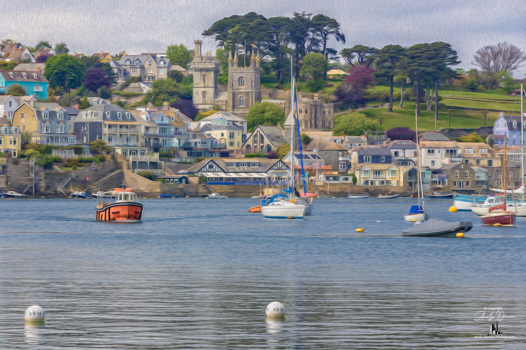 The Polruan ferry leaving Fowey.  Colourful boats moored in calm water with a hillside town featuring houses and trees in the background.