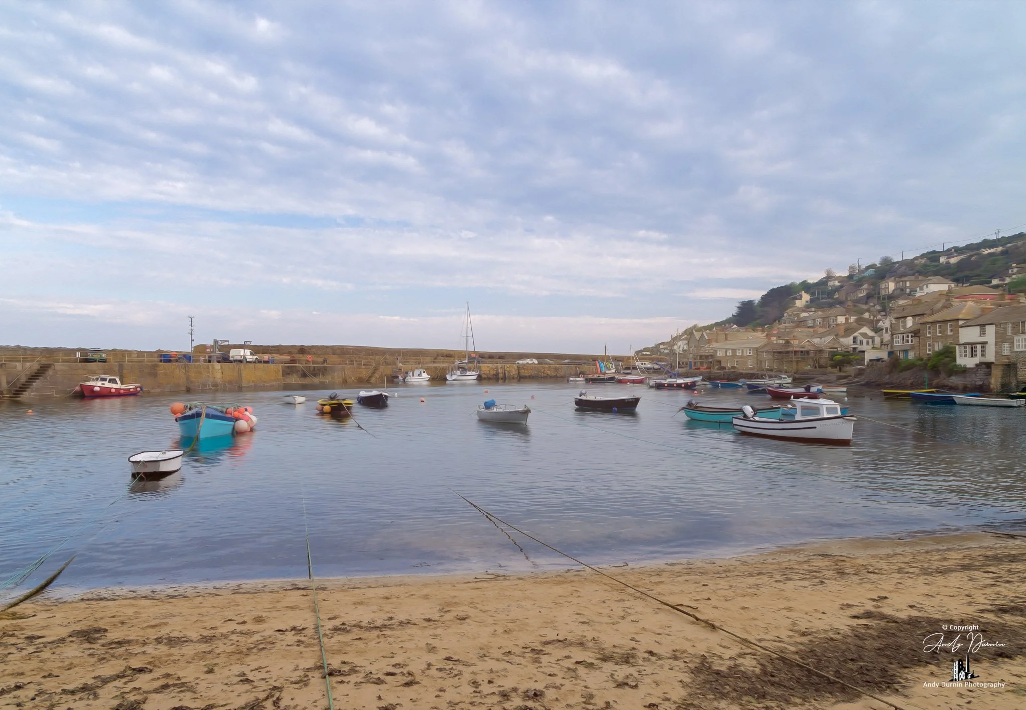 A fine art print of Mousehole Harbour in Cornwall at low tide, with sandy foreground textures, moored boats, the old harbour wall and the village climbing the hillside beyond. With its painterly finish and soft coastal light, this Cornwall wall art p