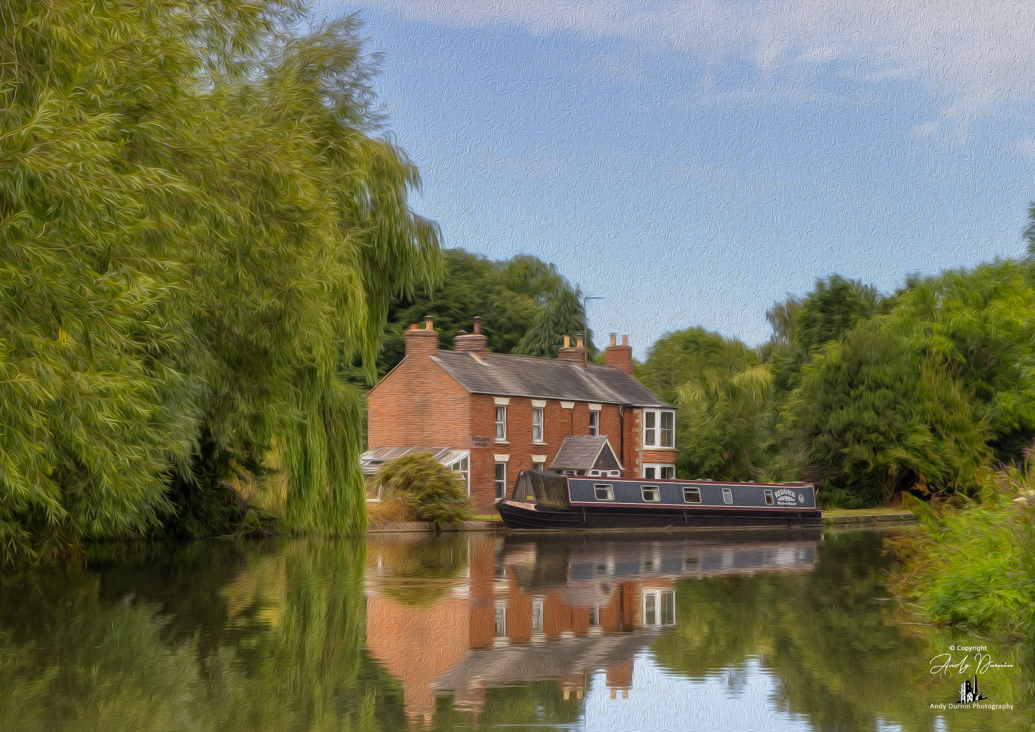 A canal scene with a brick house and a black narrowboat moored on the water, surrounded by lush green trees under a clear blue sky, with reflections in the water.