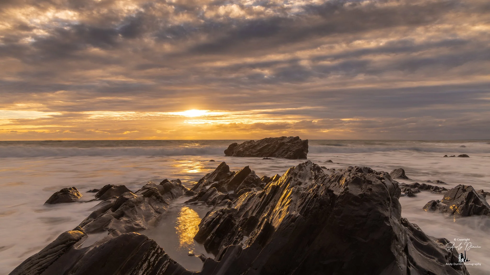 Sun setting over Dollar Cove, Cornwall with long-exposure surf, dark jagged rocks and golden reflections.