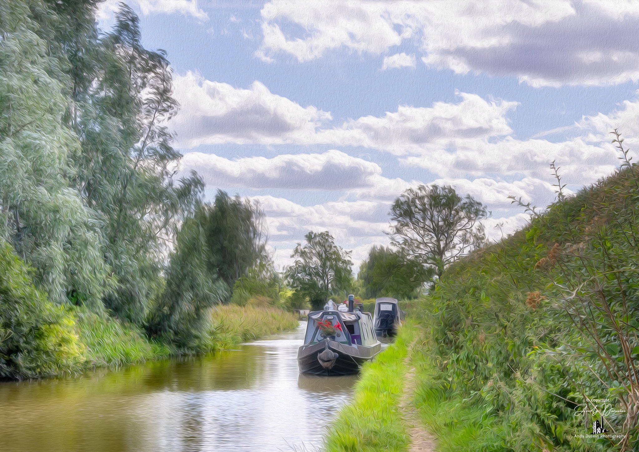 A narrowboat on the Oxford Canal moored along the bank, surrounded by green trees and bushes, with a partly cloudy sky overhead.