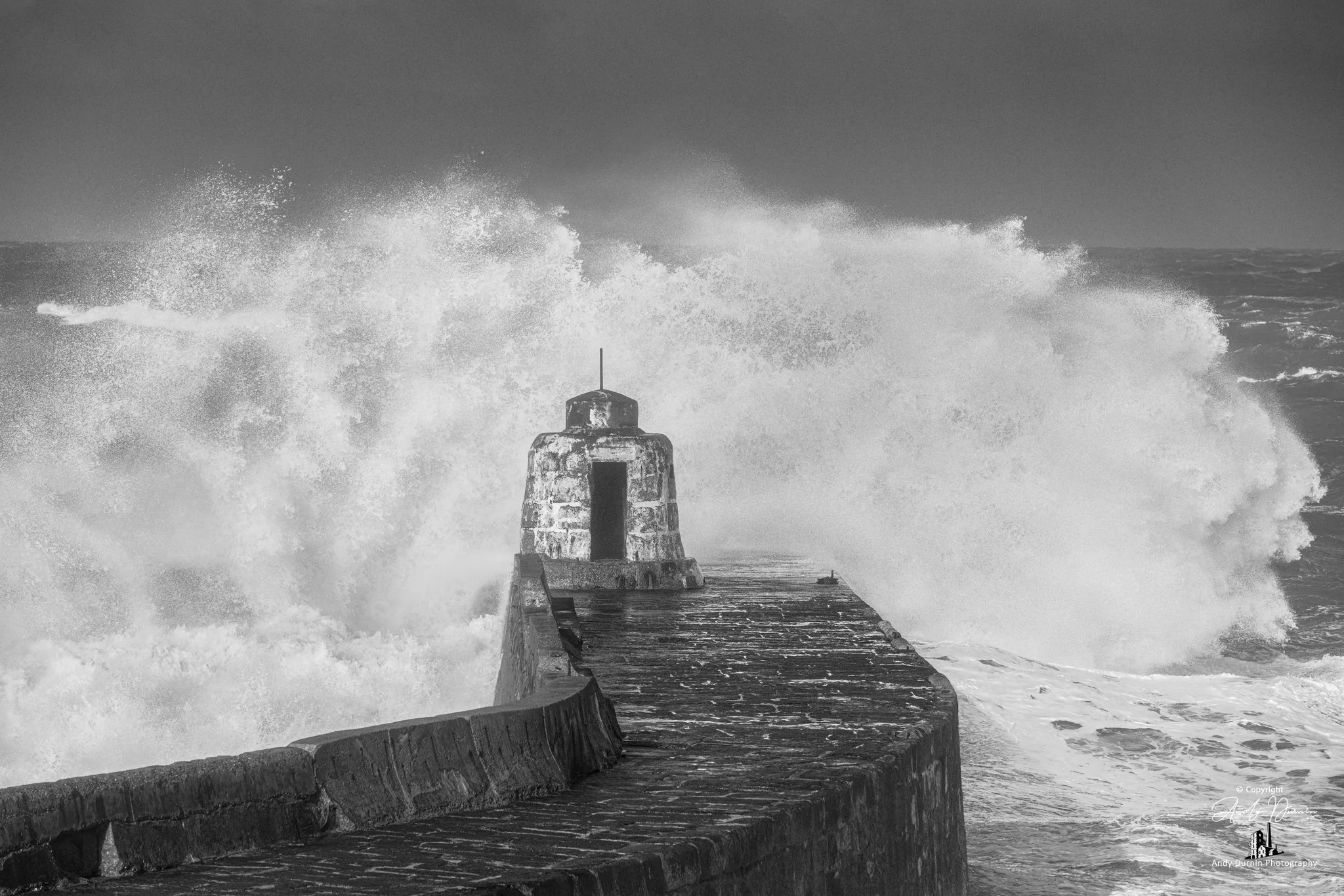 Black and white photo of a storm wave crashing over Portreath harbour wall with the Monkey Hut.