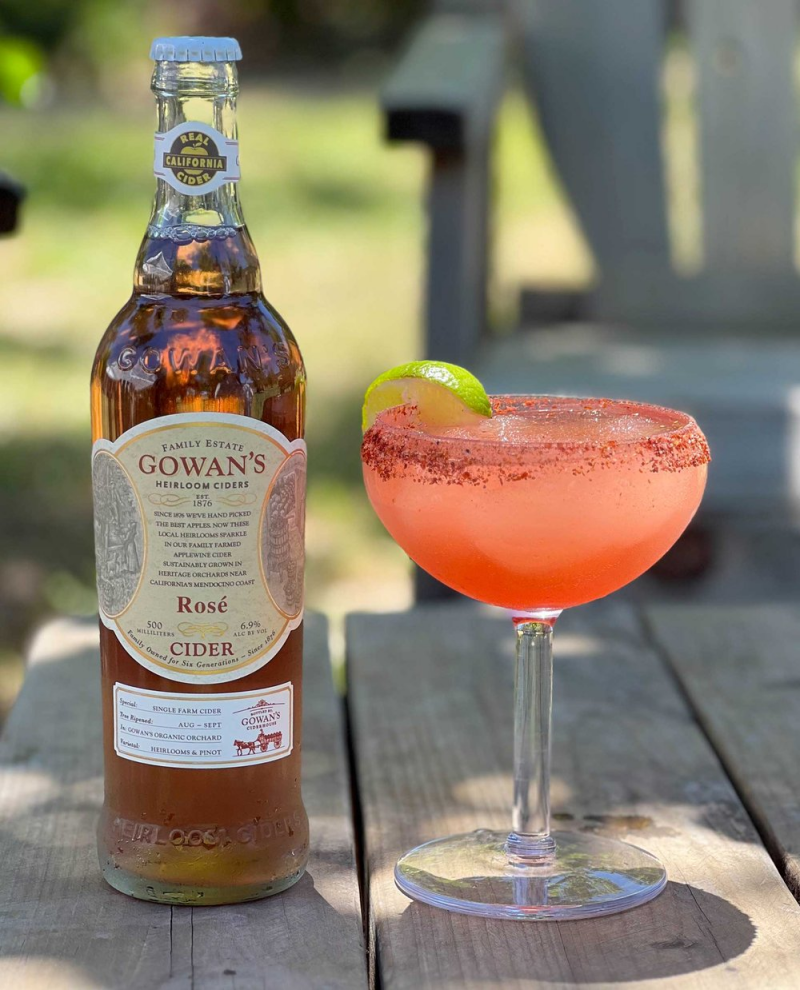 A bottle of Gowan's Heirloom Ciders Rosé sits on a wooden table next to a glass of pink cider with a lime wedge and chili powder rim, outdoors.