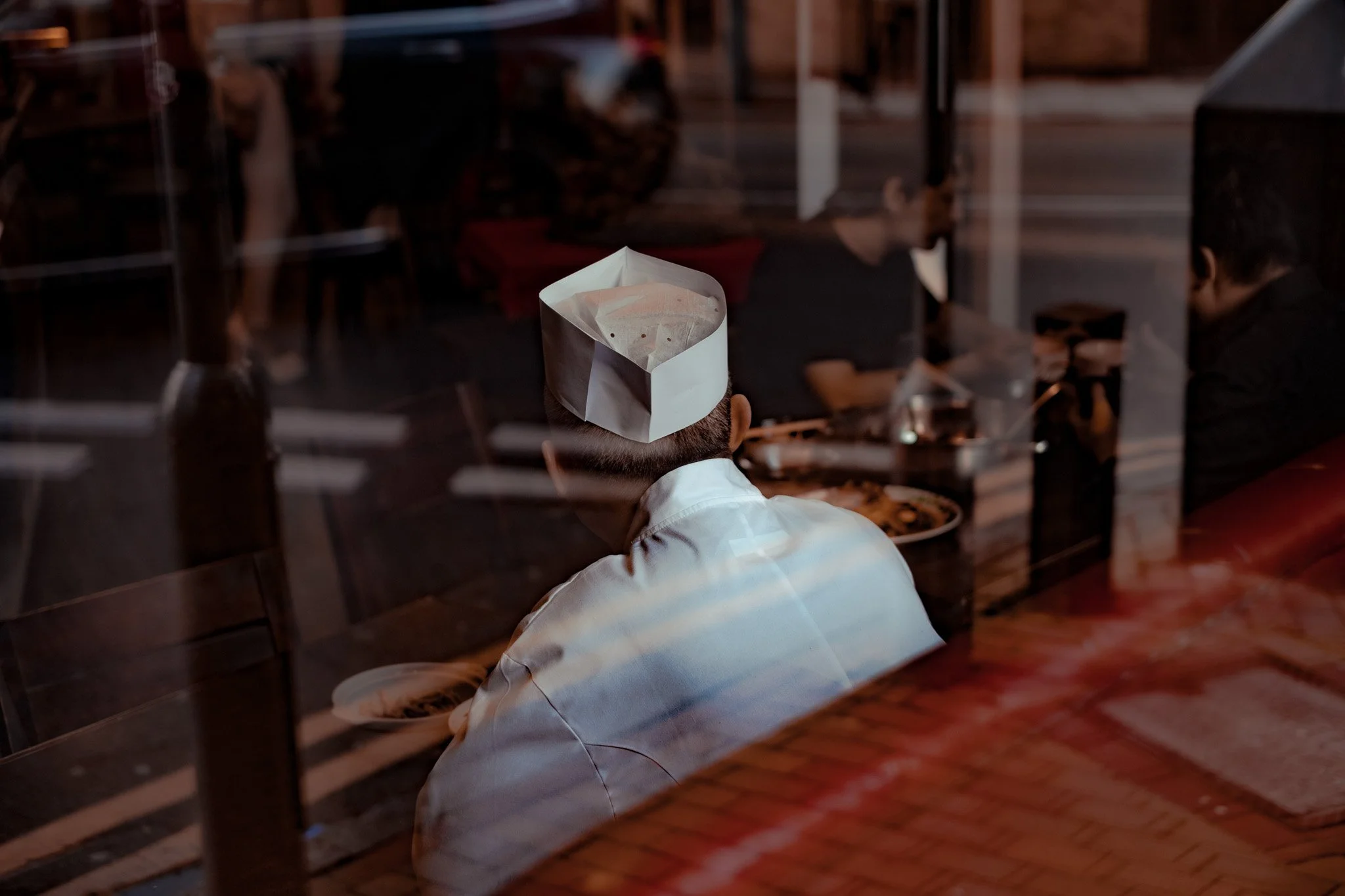 Person in a white chef's coat and paper hat sitting at a restaurant table, viewed through a glass window, with other diners and restaurant interior in the background.