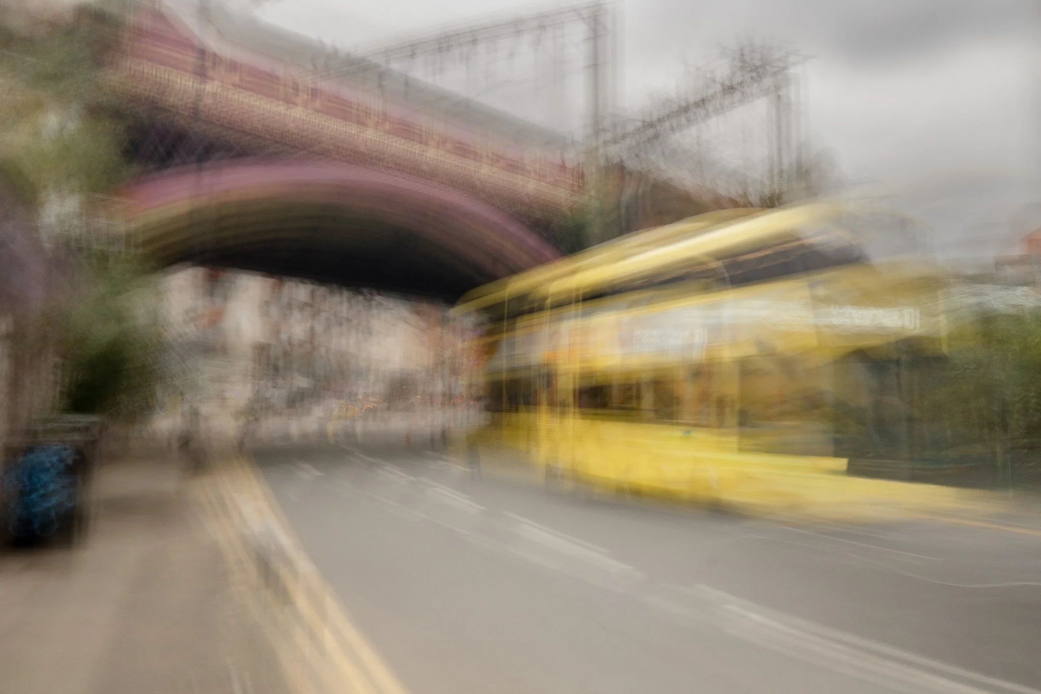 Blurred image of a yellow school bus on a gray street with an overcast sky, possibly in motion near an overpass or bridge.