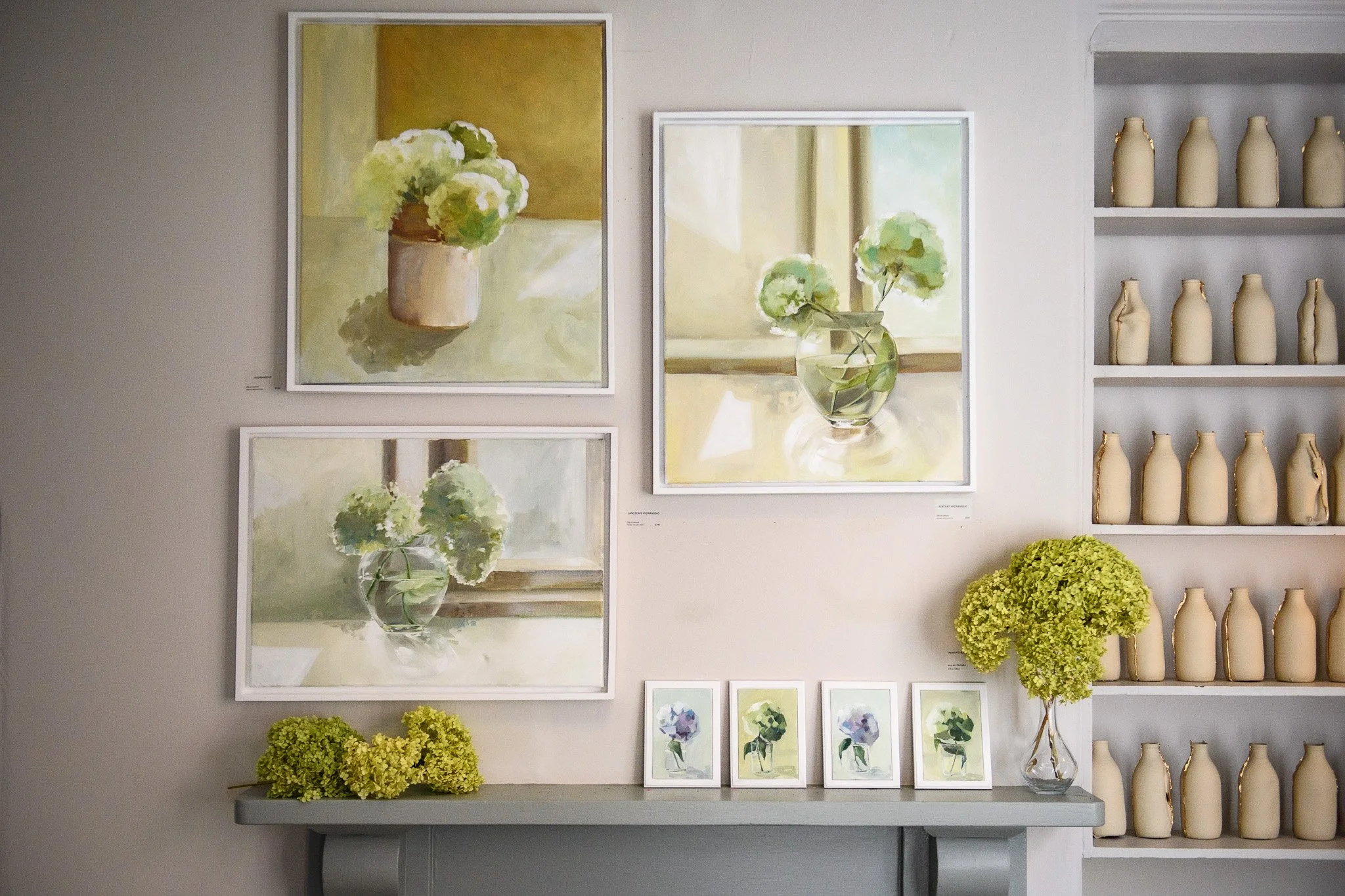 Wall display with three watercolor paintings of white hydrangeas in vases, decorative white vases on shelves to the right, and yellow-green hydrangea arrangements on a table in front.