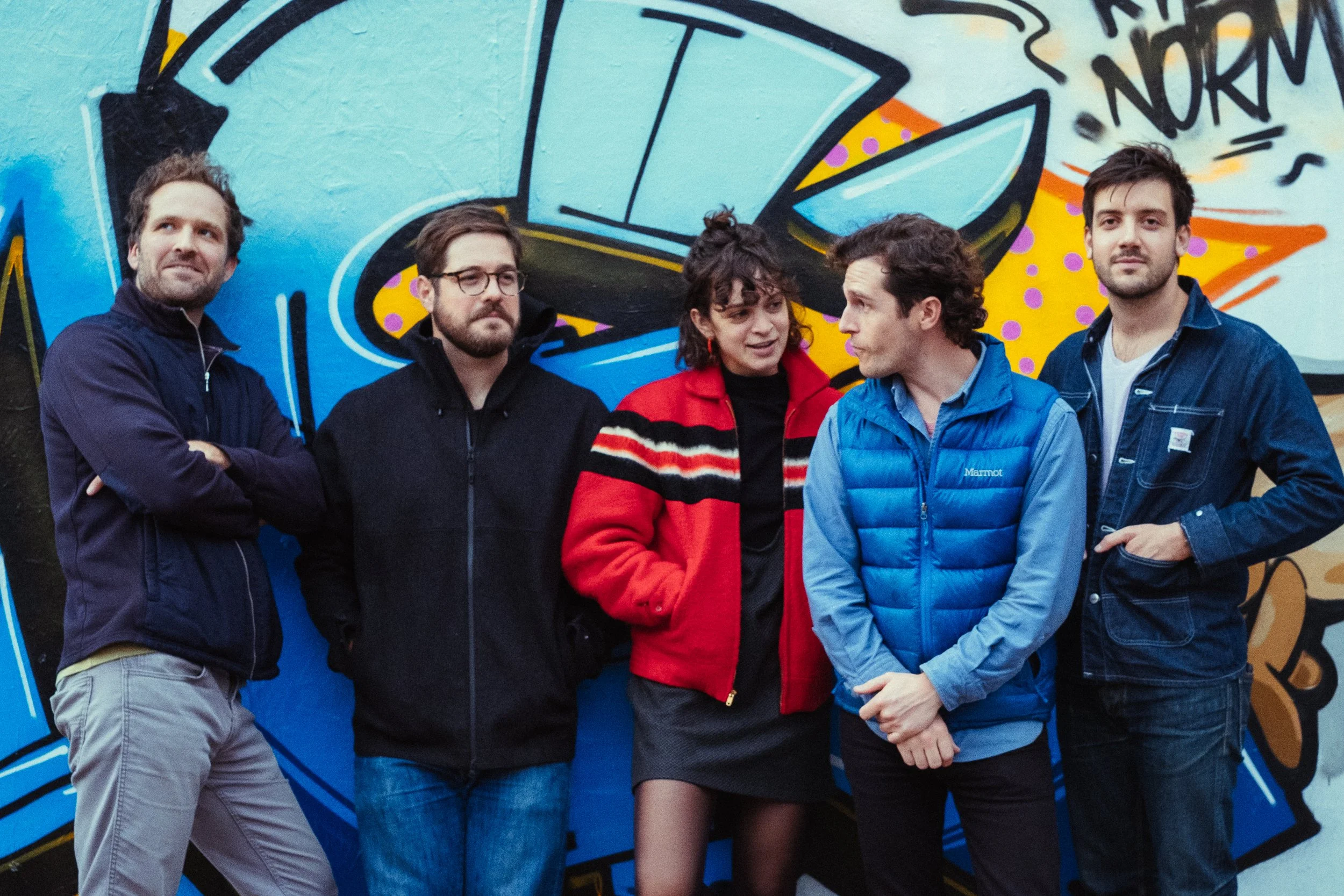 Group of five young adults standing in front of colorful graffiti mural on a wall.