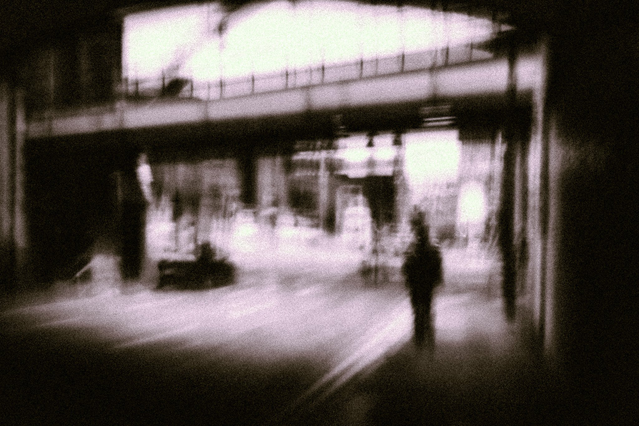 Blurred black and white photo of an outdoor scene with a bridge or overpass, several trees, and some people walking or standing.